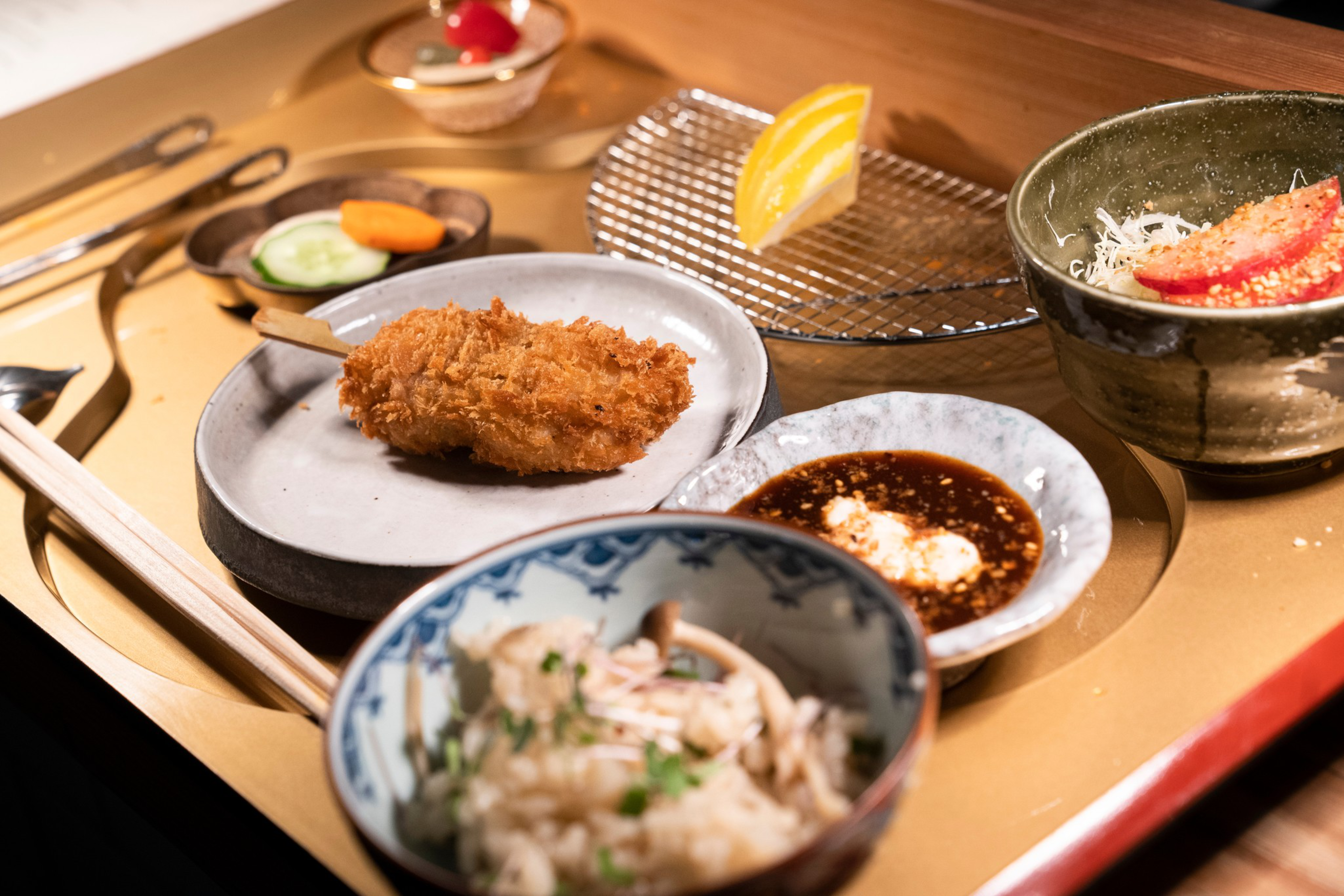 The image shows a meal with a fried item on a stick, a bowl of rice, pickled vegetables, a dipping sauce, salad with tomato and cabbage, and a lemon wedge.