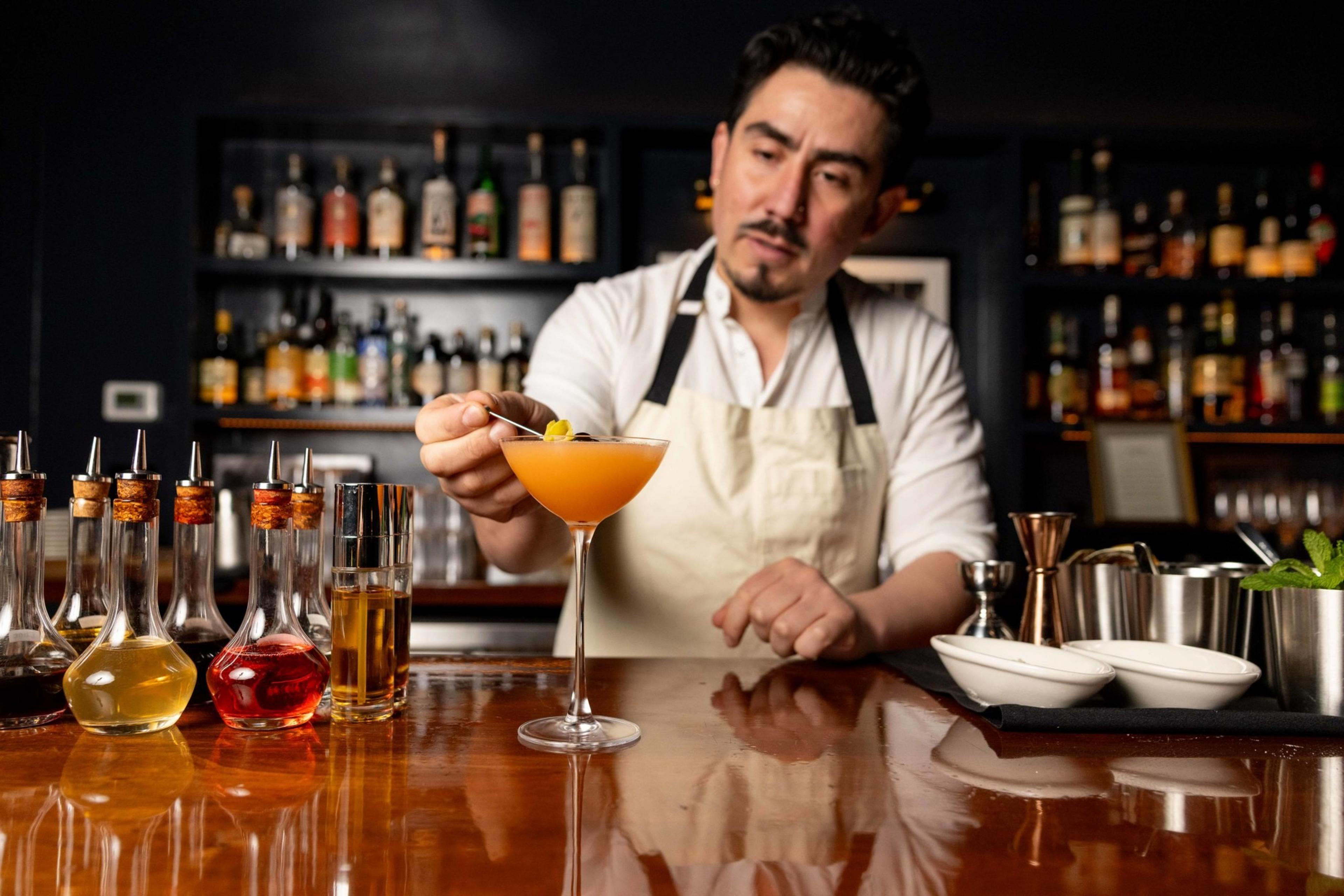 A bartender in an apron garnishes an orange cocktail with a twist in a cozy bar. Various drink ingredients and tools are neatly arranged on the counter.