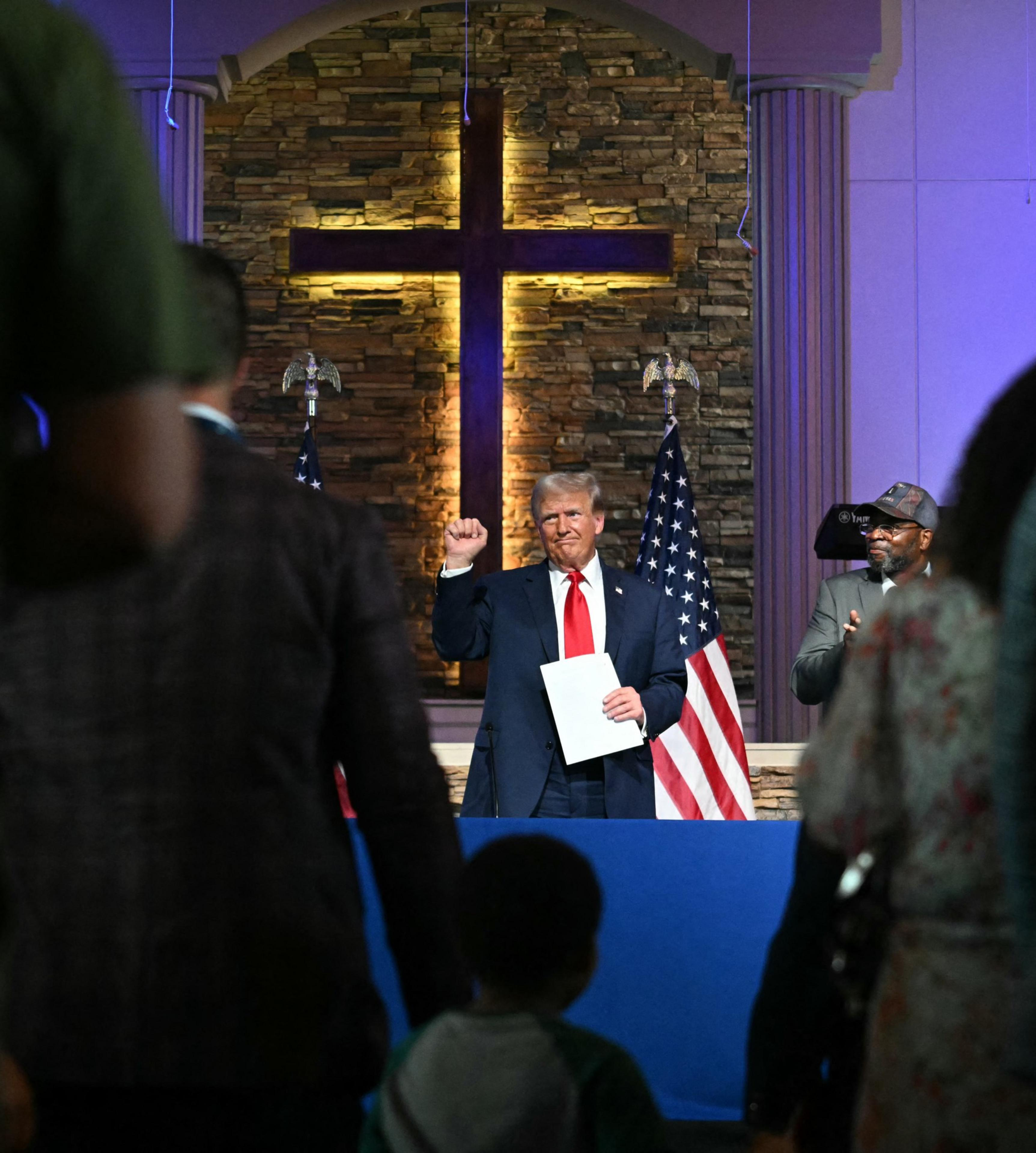 A man in a suit and red tie stands in front of a stone wall with a large illuminated cross, holding a document and raising his fist, flanked by American flags.