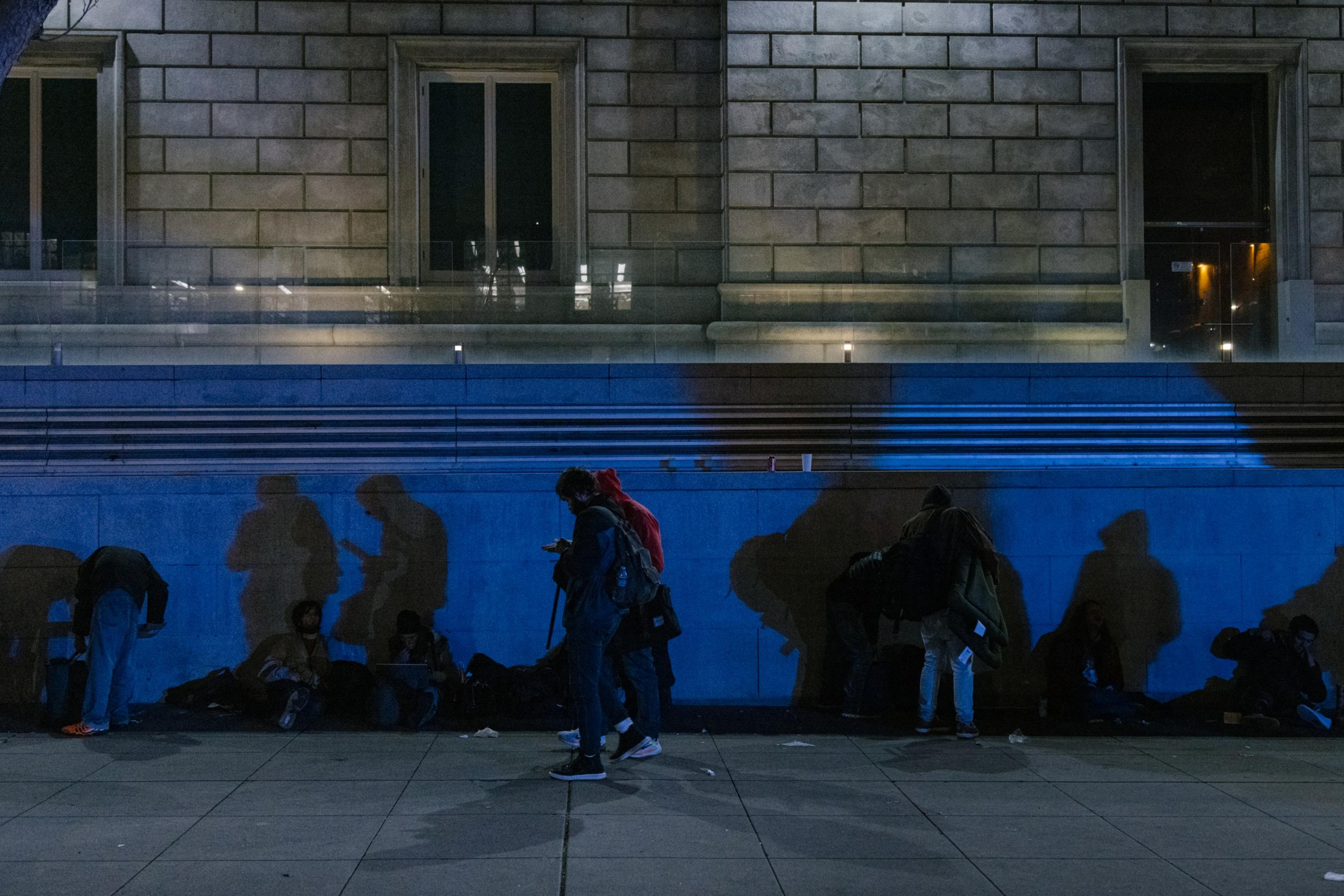 People casting shadows on a blue-lit building wall at night; some are seated, one stands using a phone.