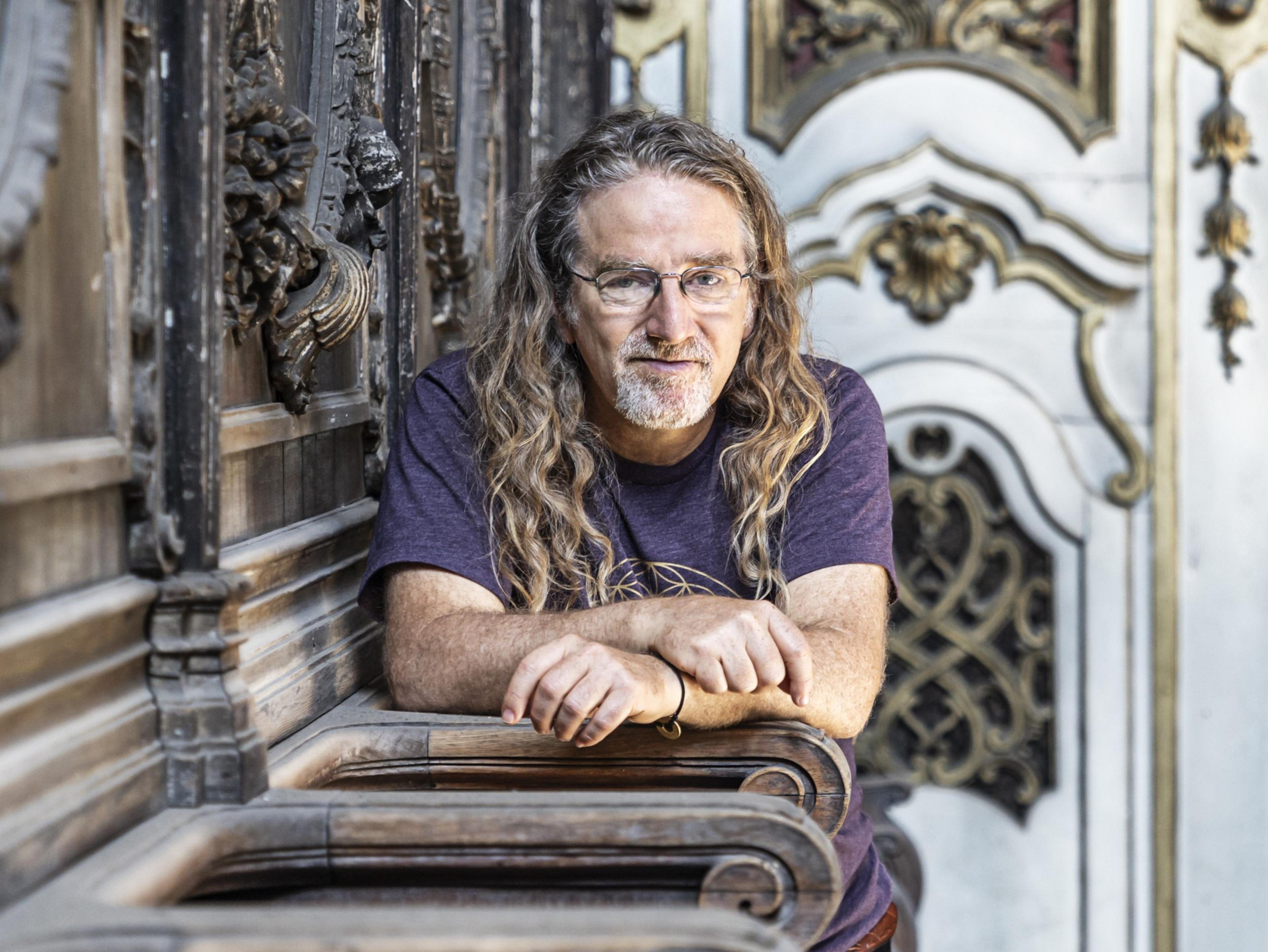 A man with long hair and glasses leans on a wooden railing in an ornate room, featuring detailed carvings and intricate designs on the walls.