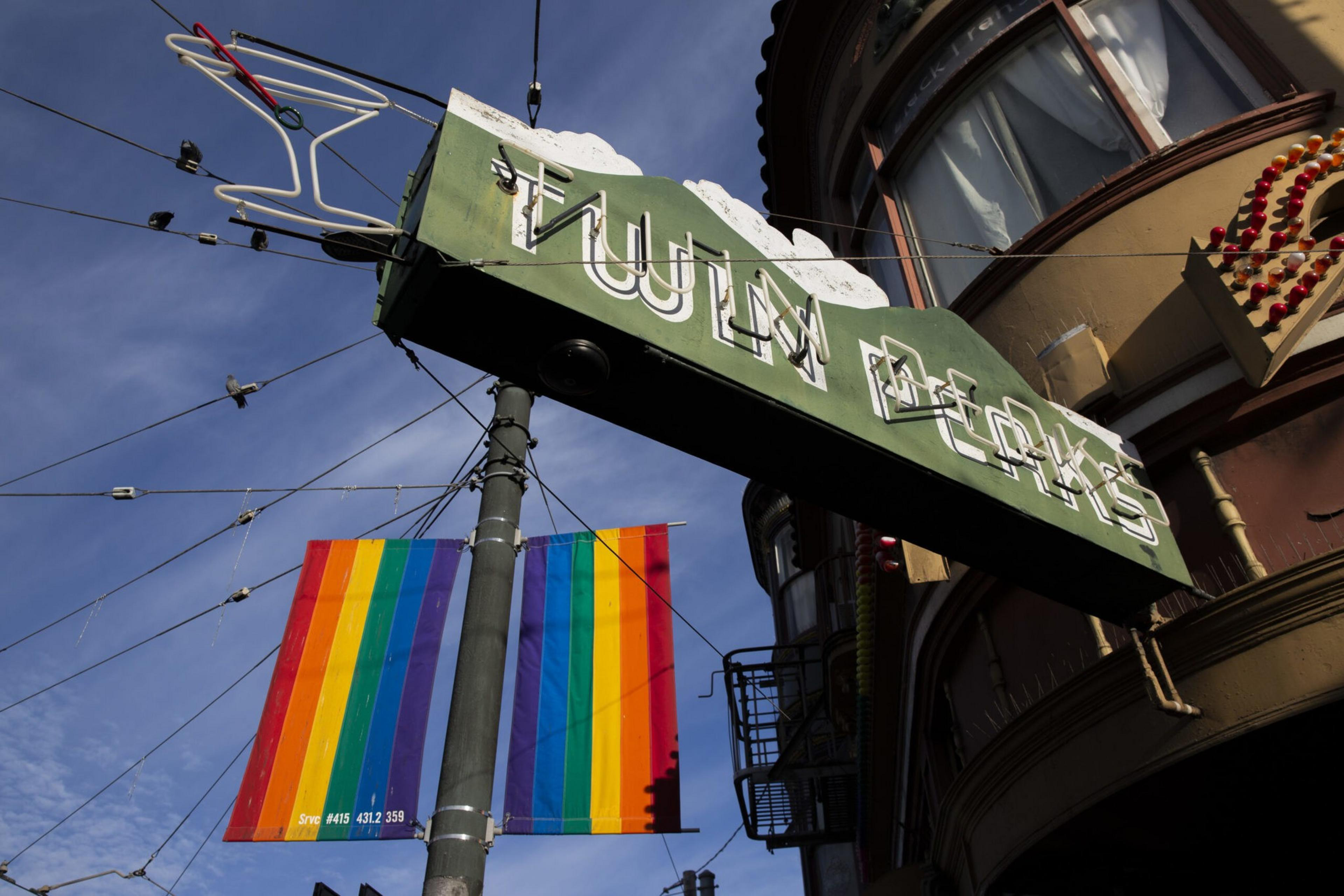 A green sign with white neon letters hangs on a pole against a blue sky. Below it, a vibrant rainbow flag is displayed.