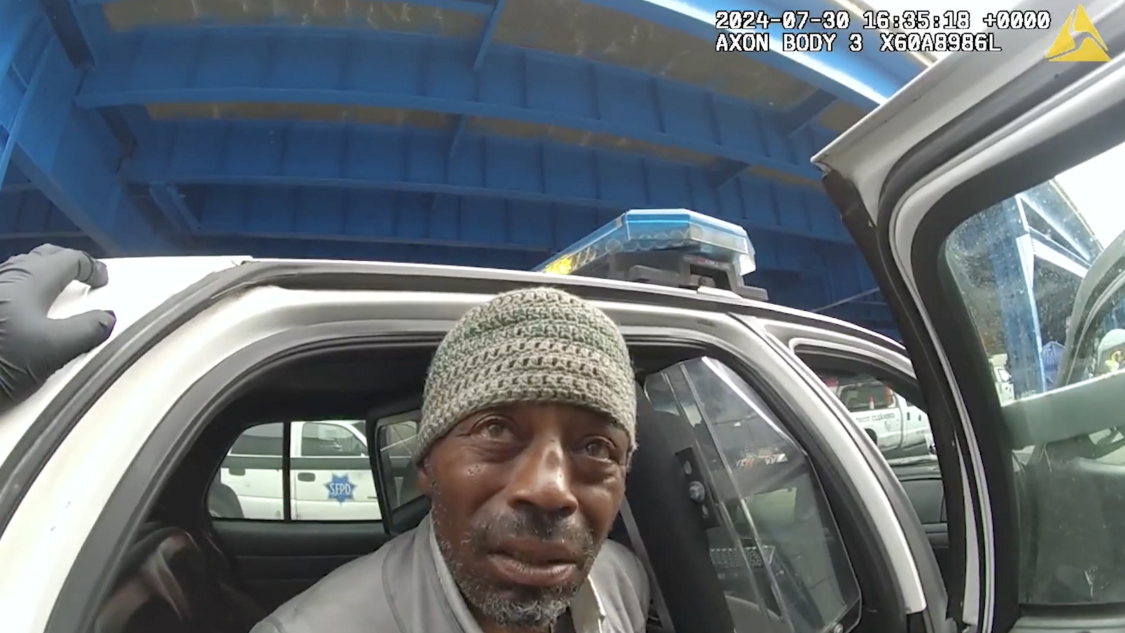 A man wearing a knit cap sits in the back of a police car with the door open. A gloved hand appears on the door frame, and a blue structure is visible above.