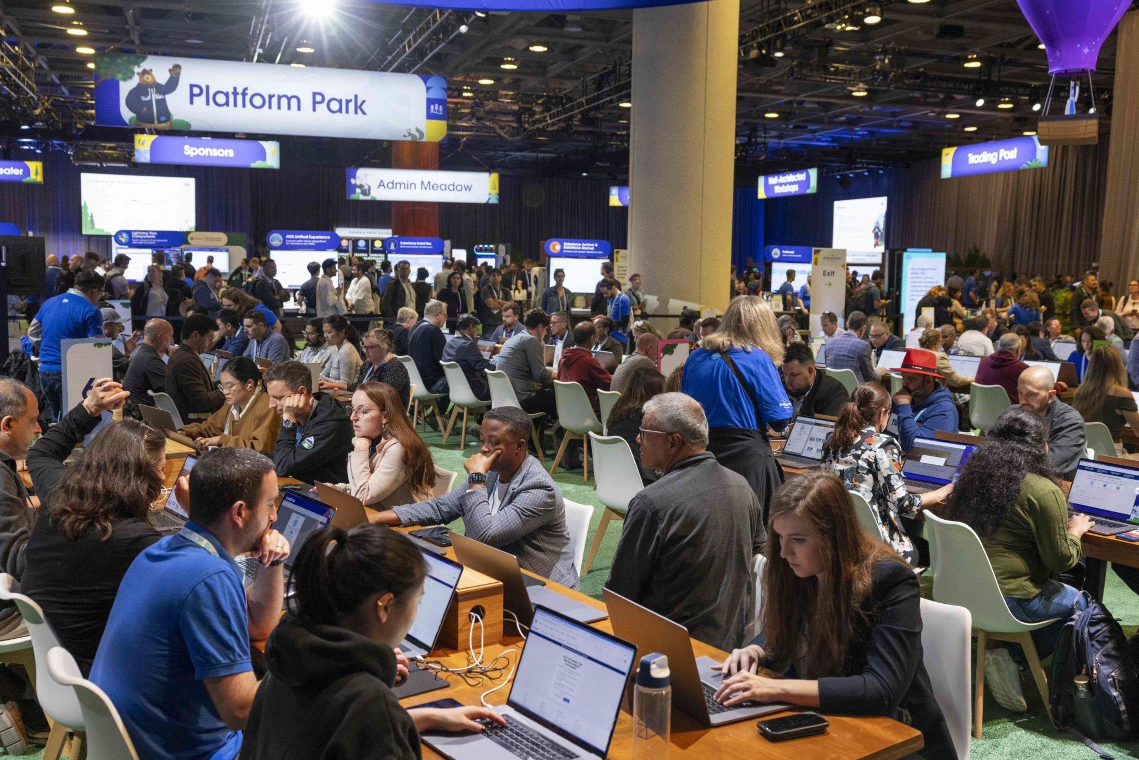People fill a tech conference hall, working on laptops at long tables under signs reading &quot;Platform Park&quot; and &quot;Admin Meadow,&quot; and other designated areas.