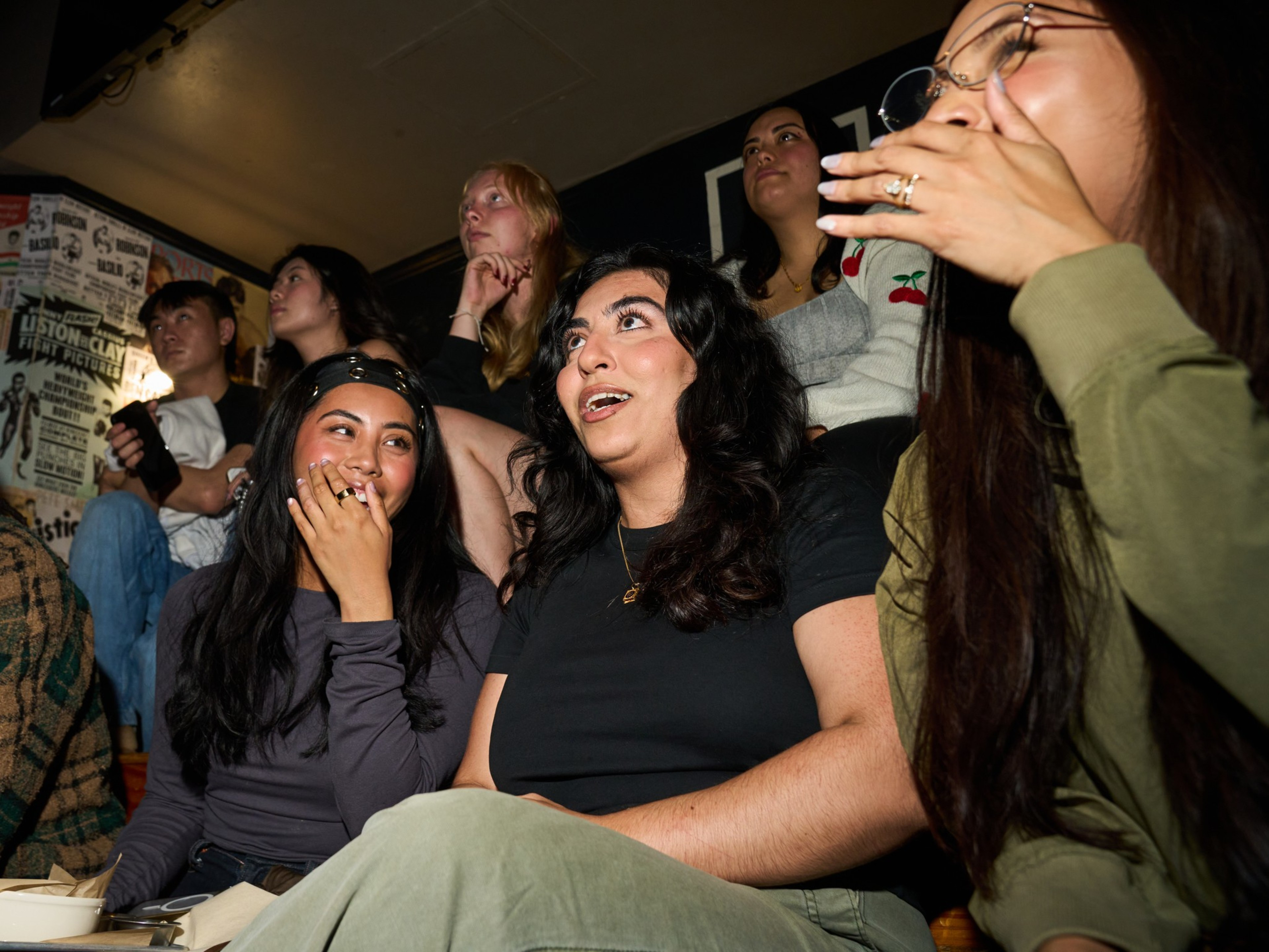 A group of people in a cozy, dimly-lit space are laughing and reacting to something off-camera. They are seated closely, with posters visible on the wall behind them.