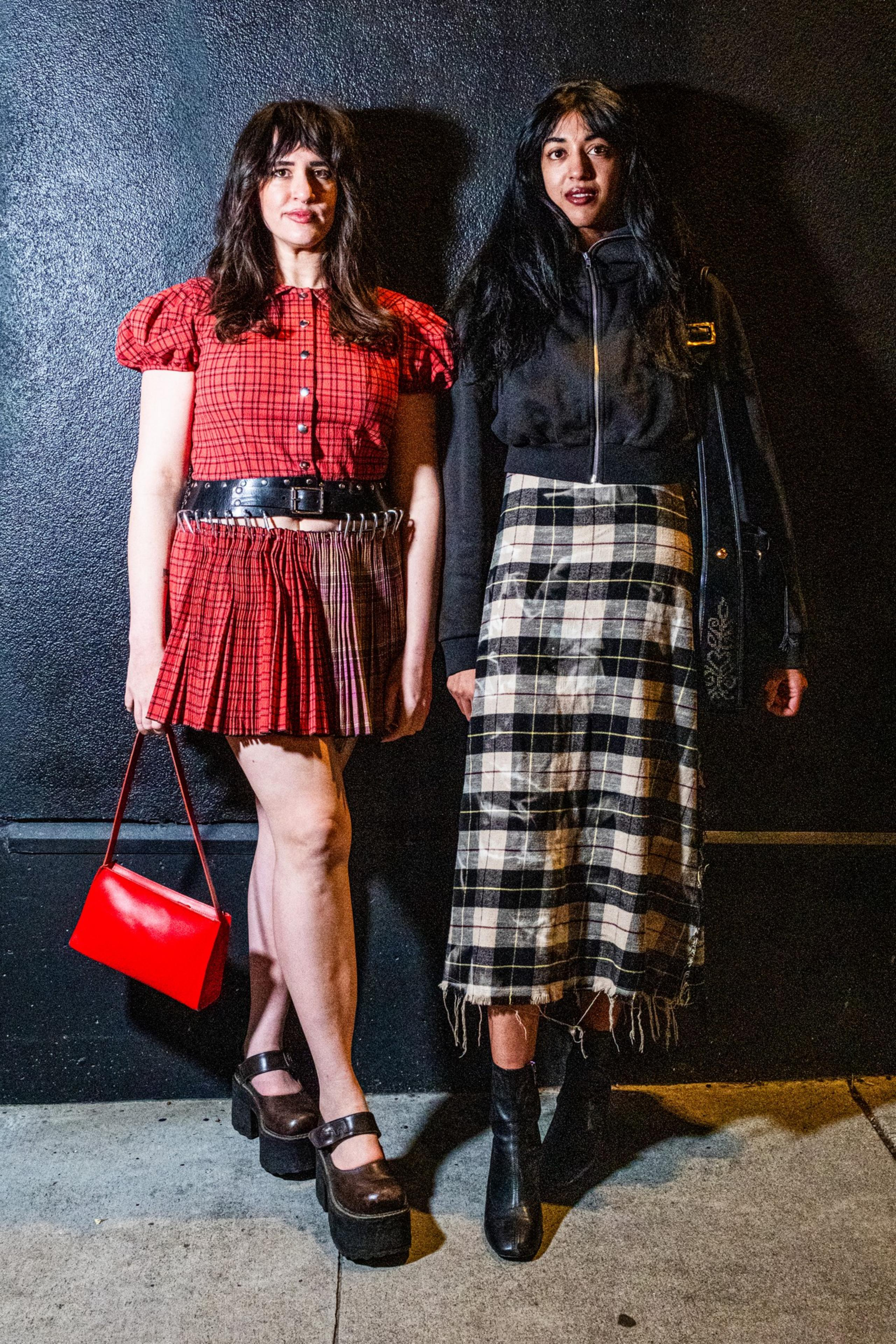 Two people stand against a dark wall. One wears a red plaid outfit and holds a red purse, the other wears a black jacket with a long plaid skirt.