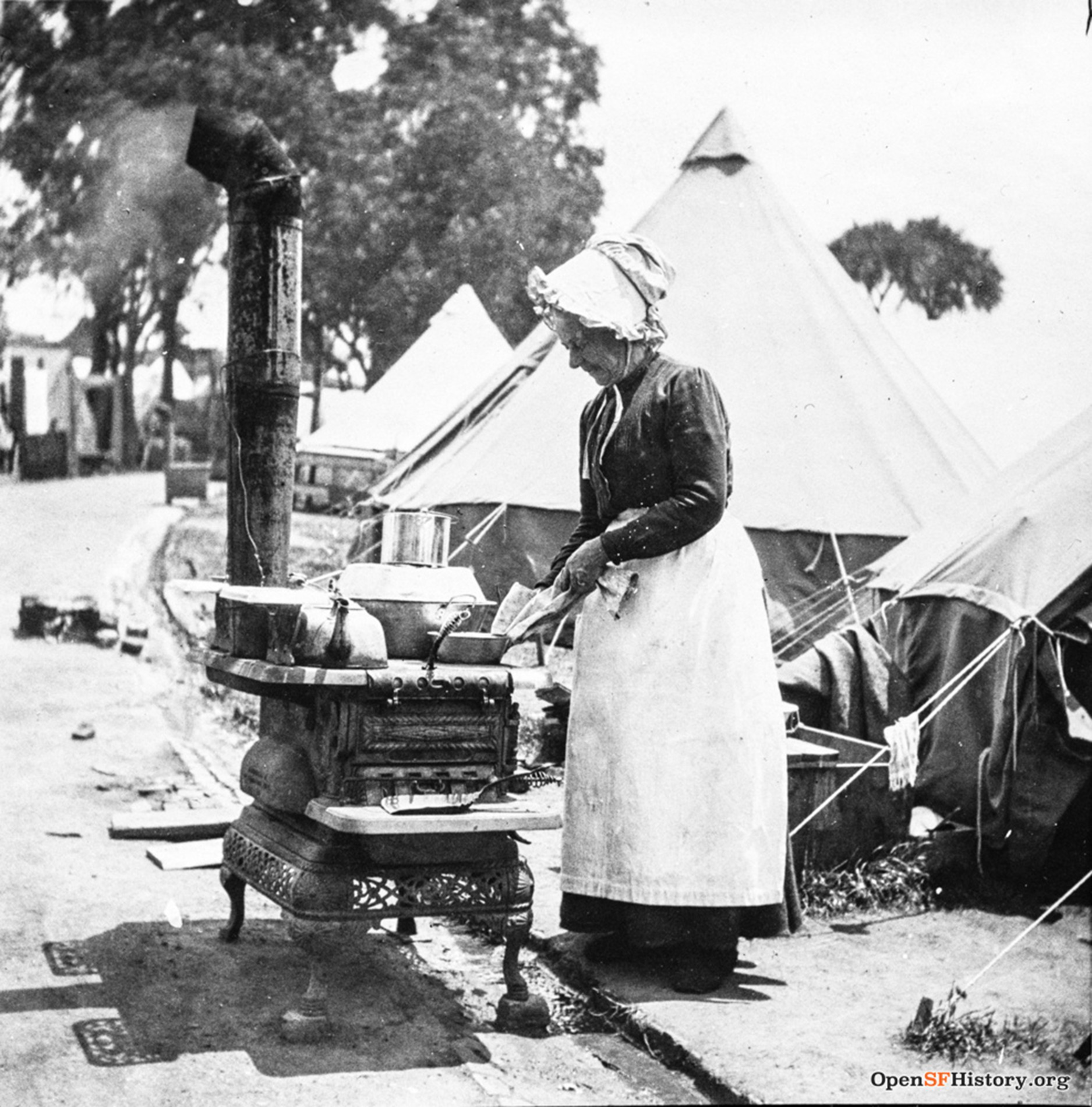 An elderly woman wearing a bonnet and apron cooks outdoors on a wood stove beside canvas tents in a camp setting.