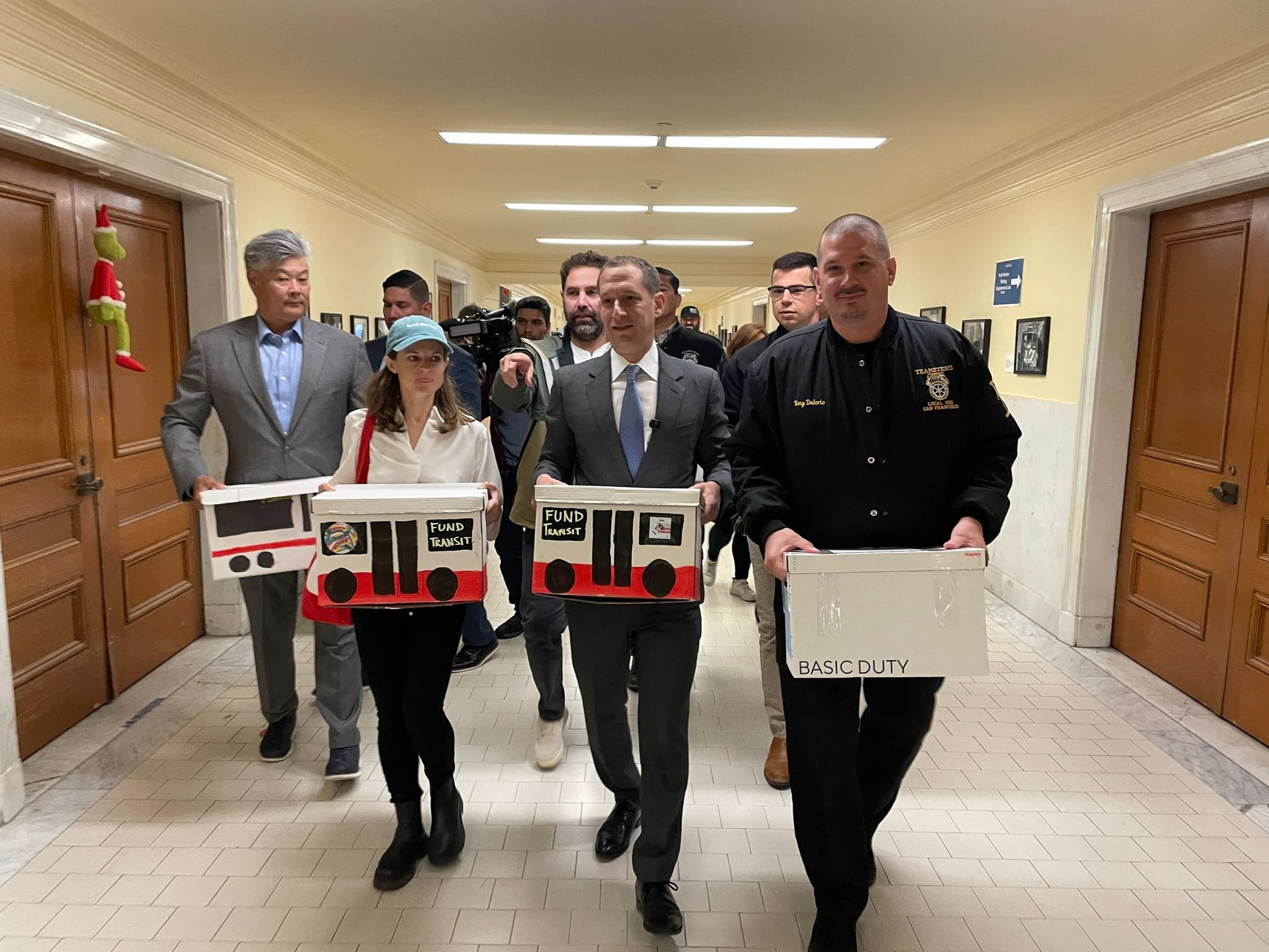 A group of people walk down a hallway, some holding boxes labeled “FUND TRANSIT” designed like buses and one box labeled “BASIC DUTY.”