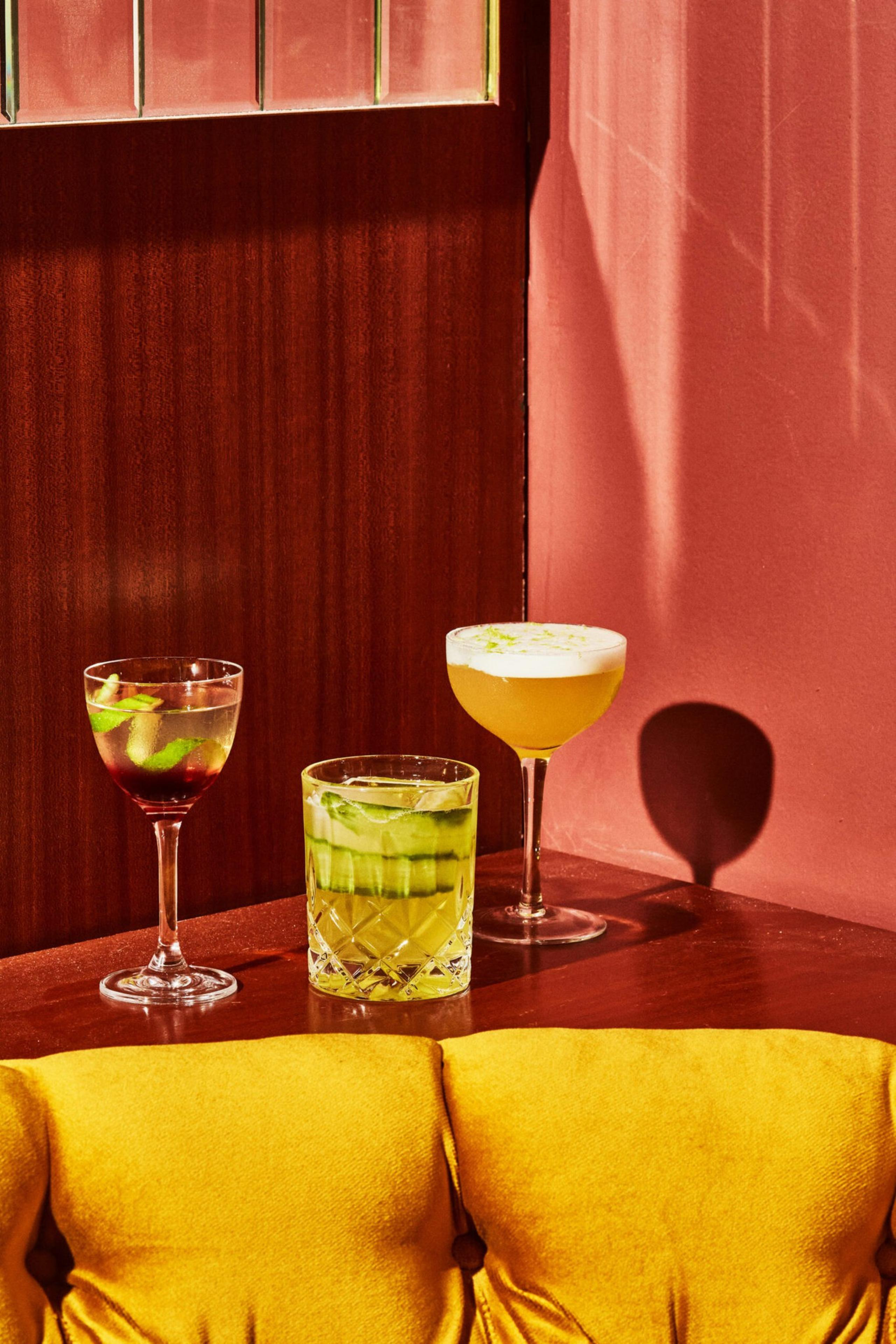 Three cocktails—one red with lime, one green with cucumber, and one frothy yellow—are arranged on a wooden surface by a pink wall and yellow cushion.