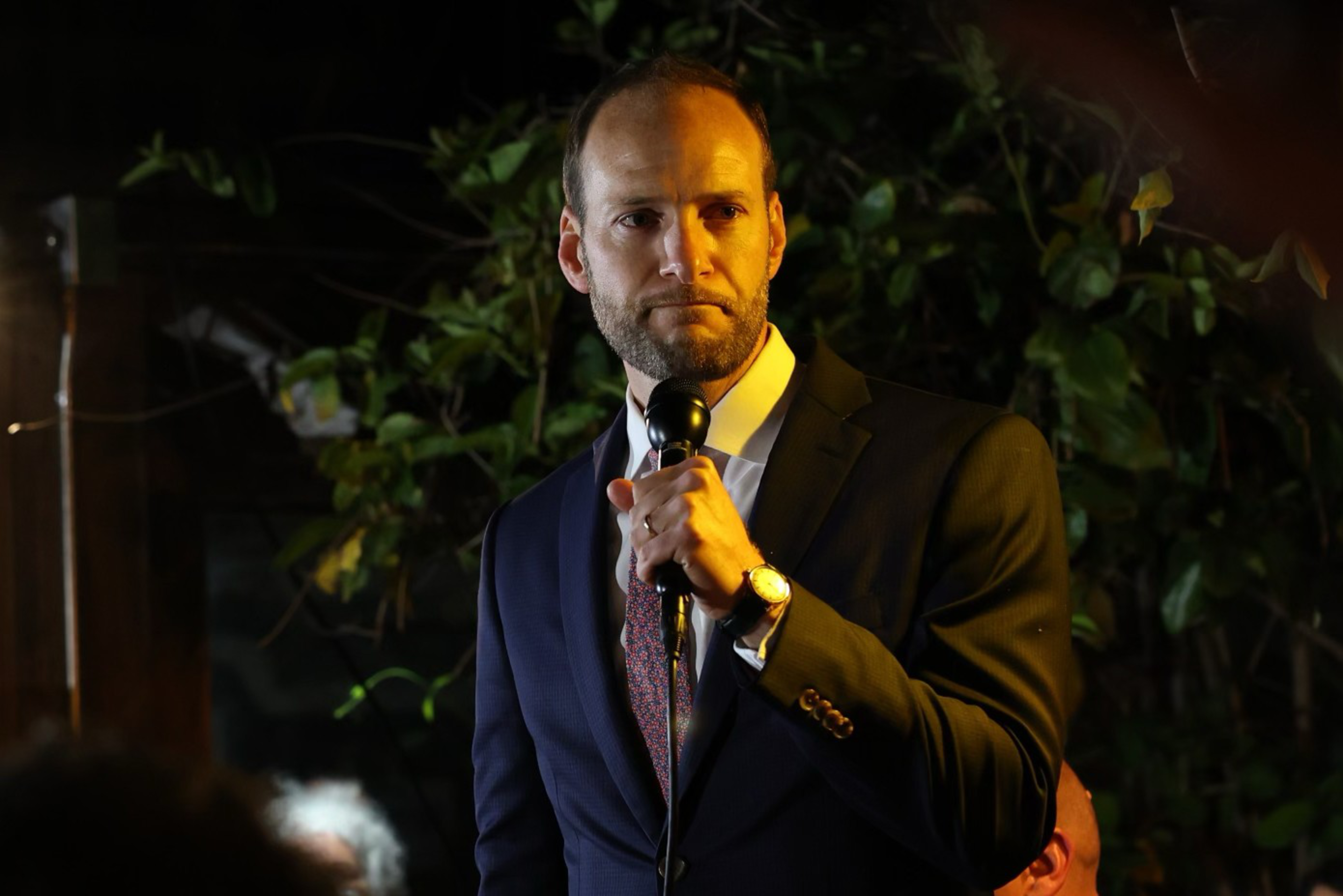 Chesa Boudin in a dark suit and tie holds a microphone, standing under warm lighting, with leafy green plants in the background. He appears to be speaking or addressing an audience.