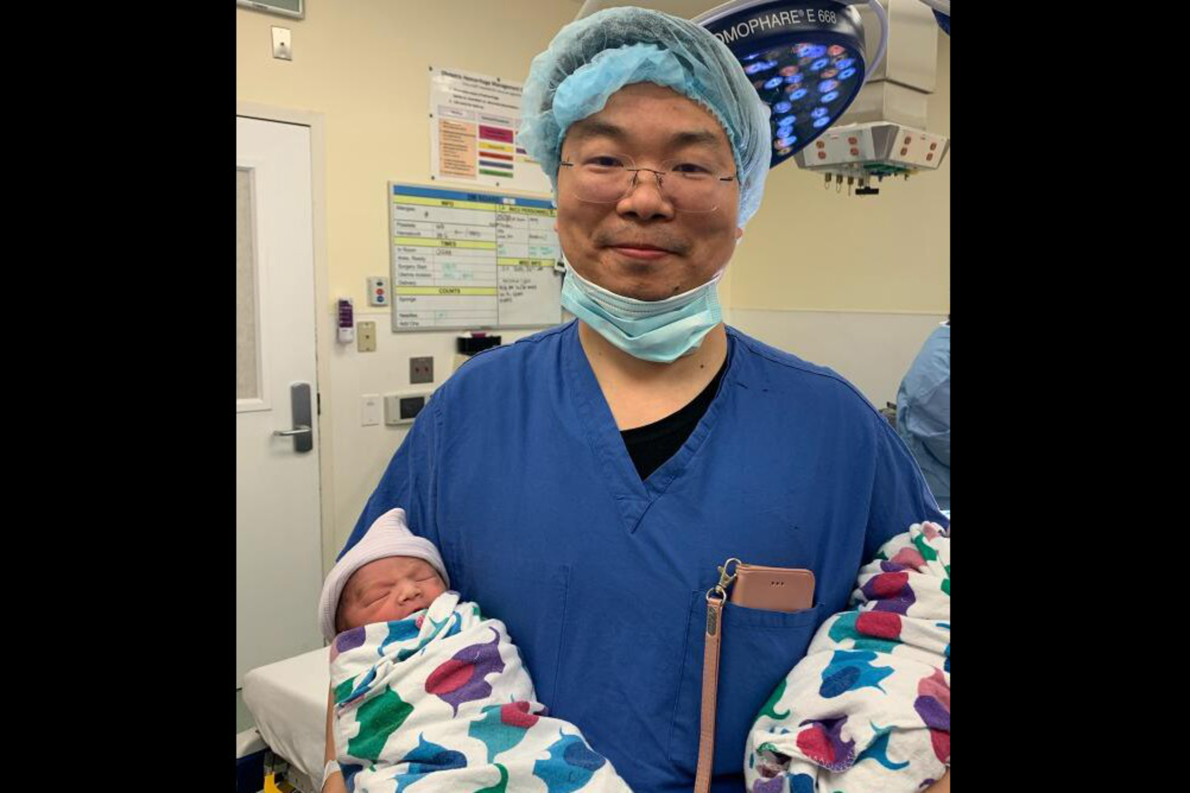 A person in blue scrubs, a hair net, and glasses gently holds two newborn babies wrapped in colorful blankets inside a hospital room.