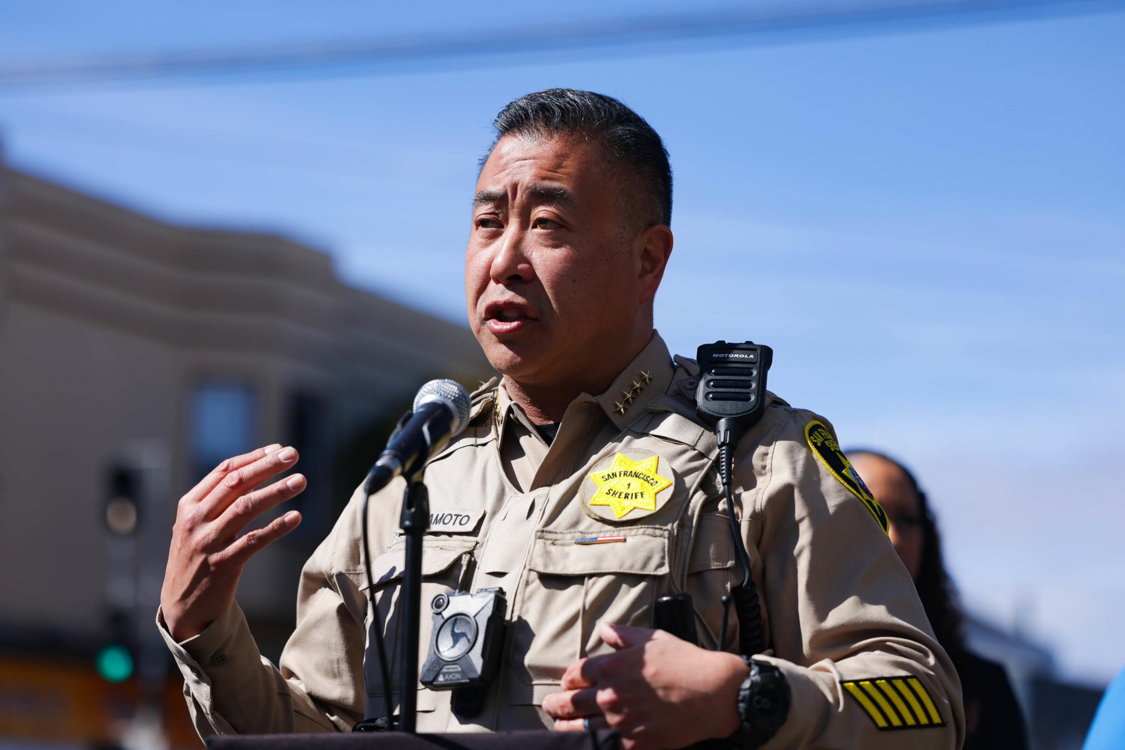 Sheriff Paul Miyamoto stands outside in front a microphone, speaking.