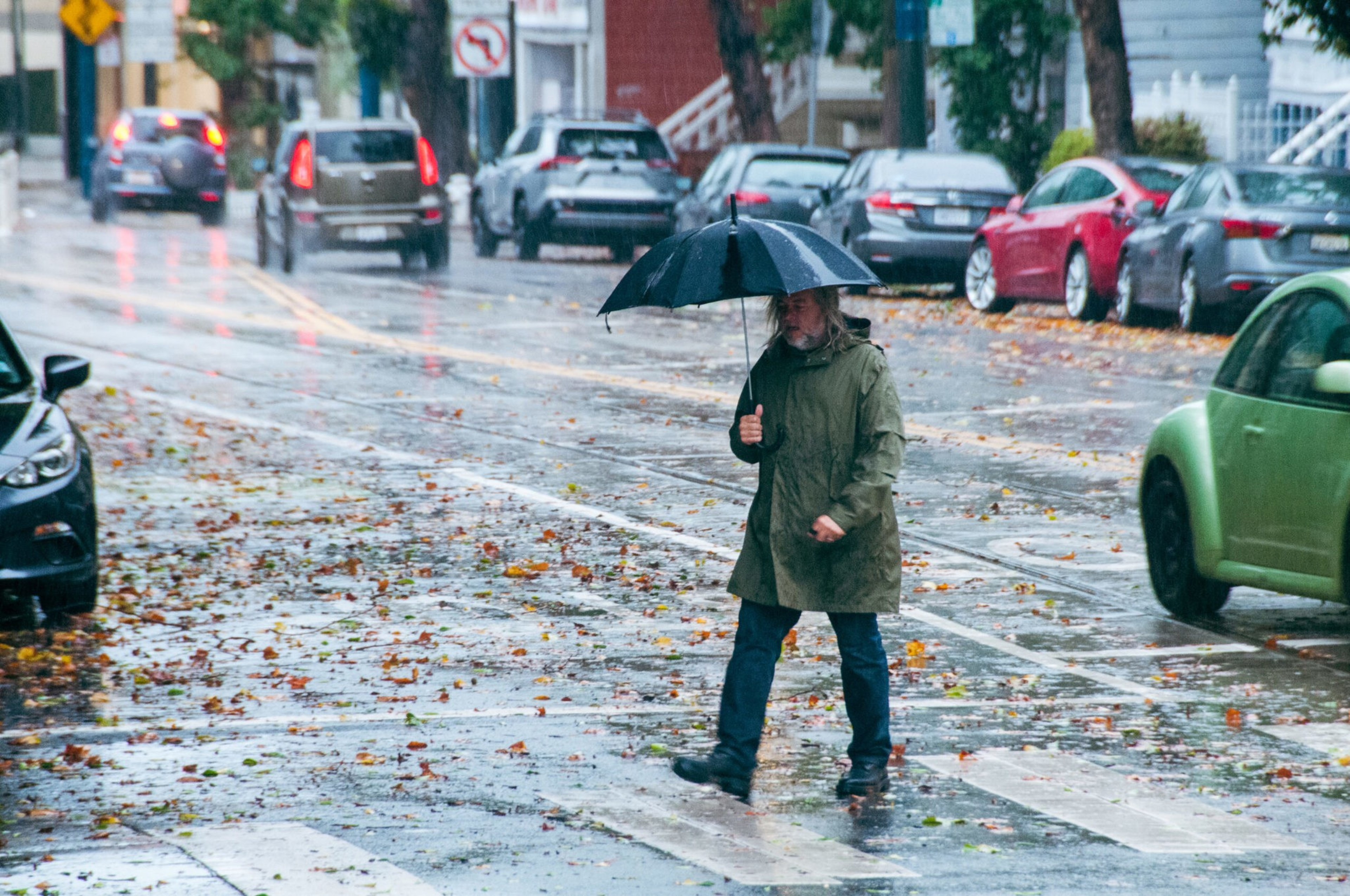 A person wearing a green coat holds a black umbrella while walking across a wet street scattered with autumn leaves on a rainy day.