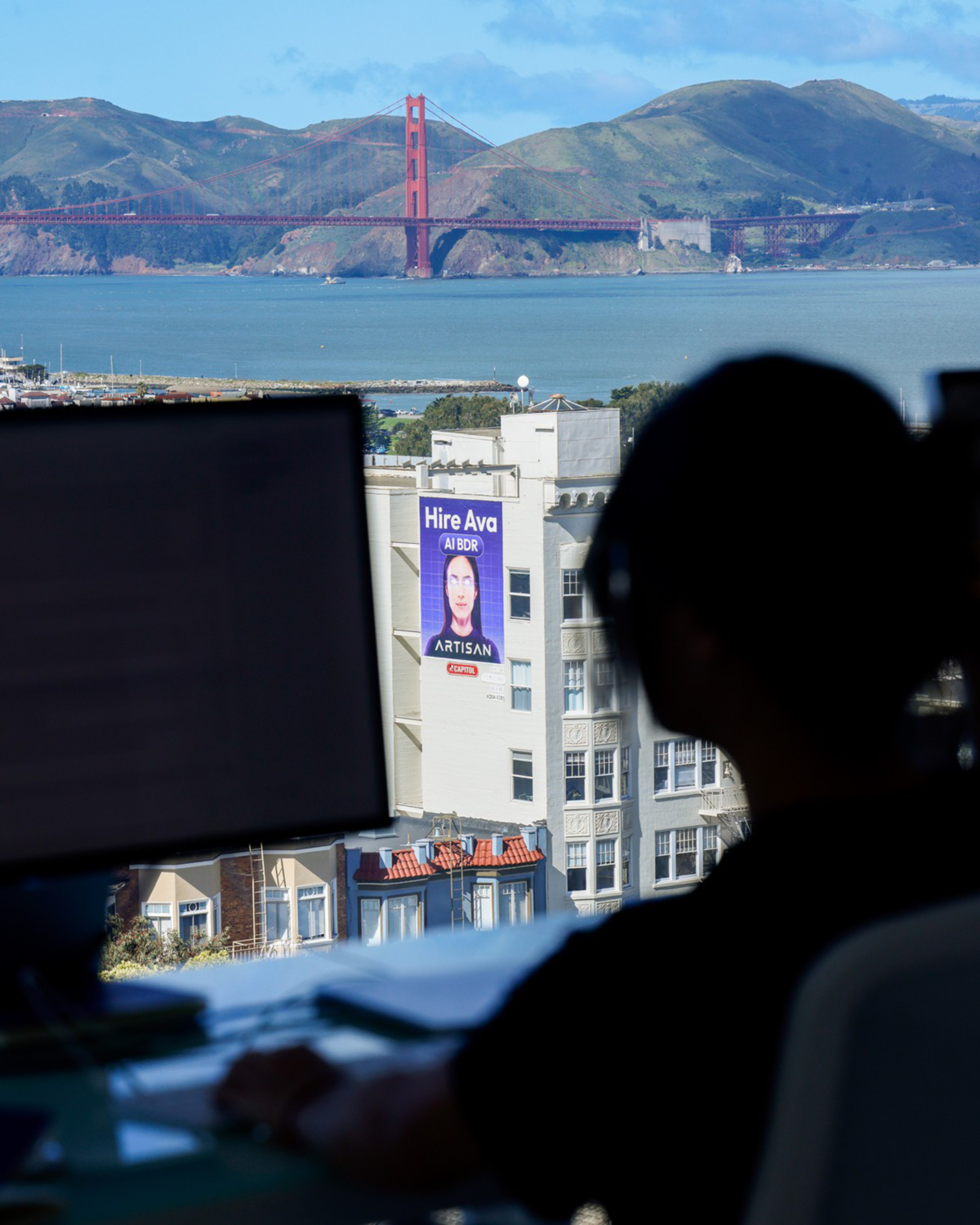 A person works at a computer with a view of the Golden Gate Bridge and San Francisco buildings. A billboard on a building advertises &quot;Hire Ava AI BDR.&quot;