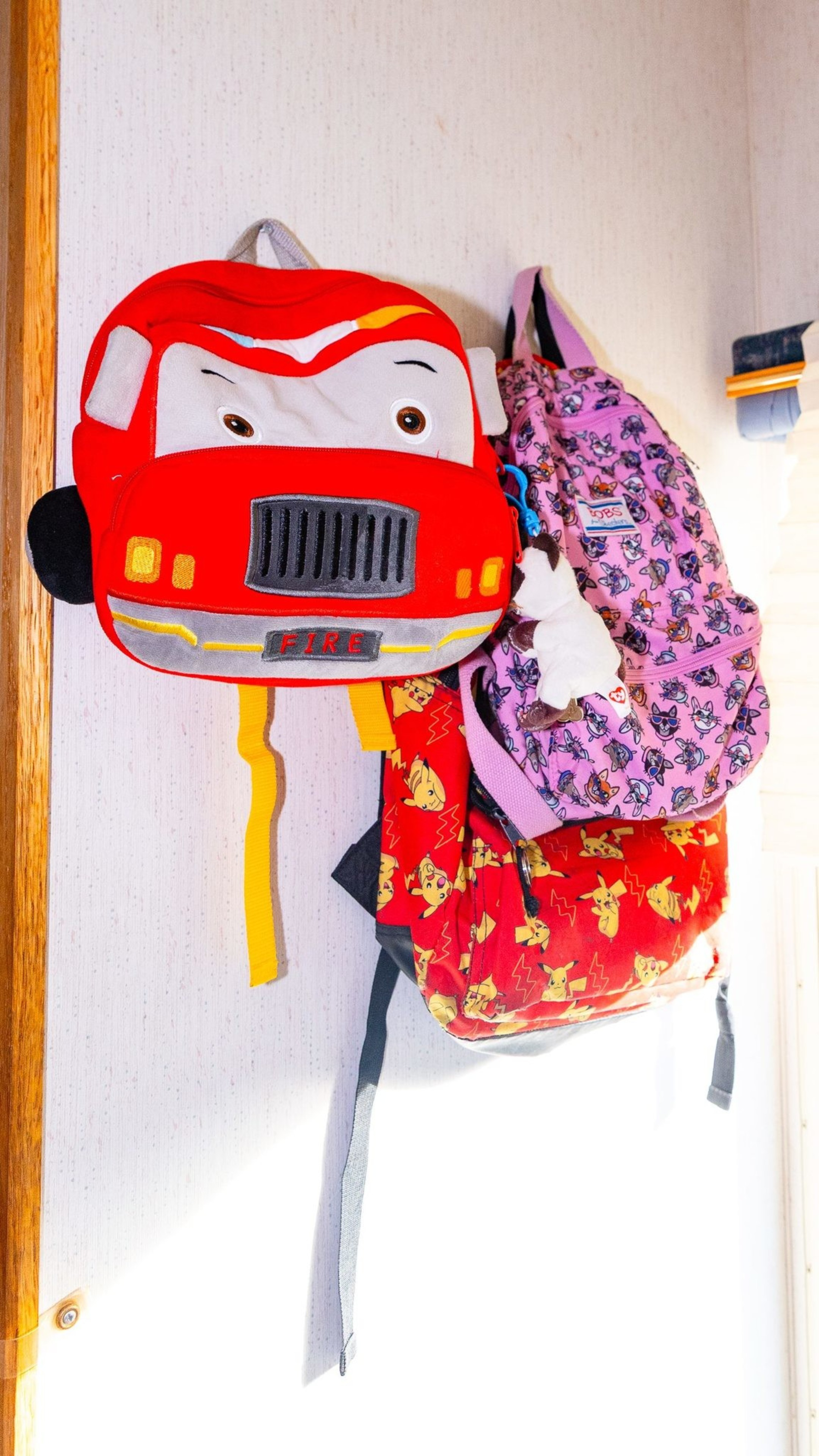 Three colorful backpacks hang on a wall: a red fire truck design, a pink one with cats, and a red one featuring Pikachu.