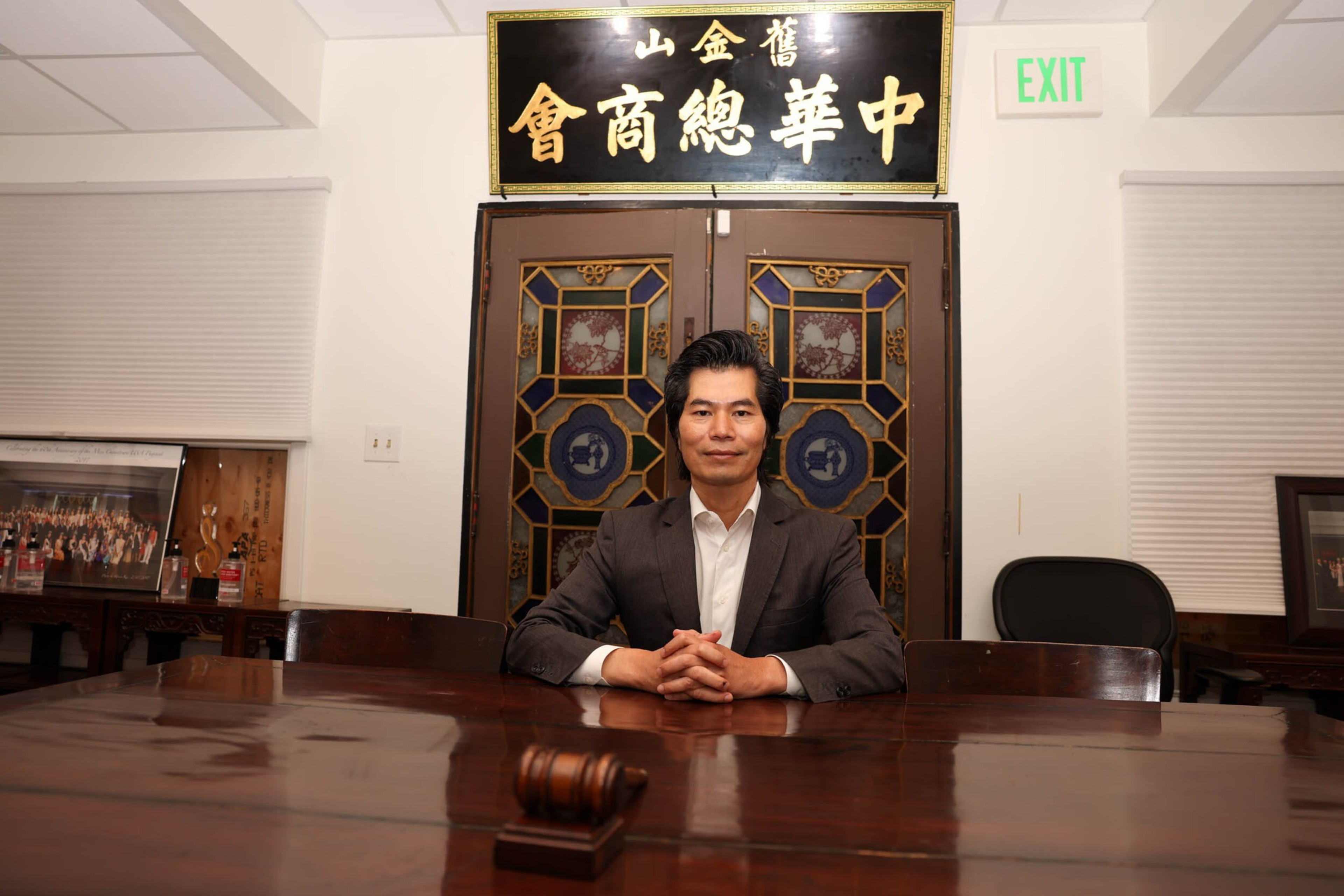 A man in a suit sits at a wooden table with a gavel, in front of ornate double doors and a black sign with gold Chinese characters above.