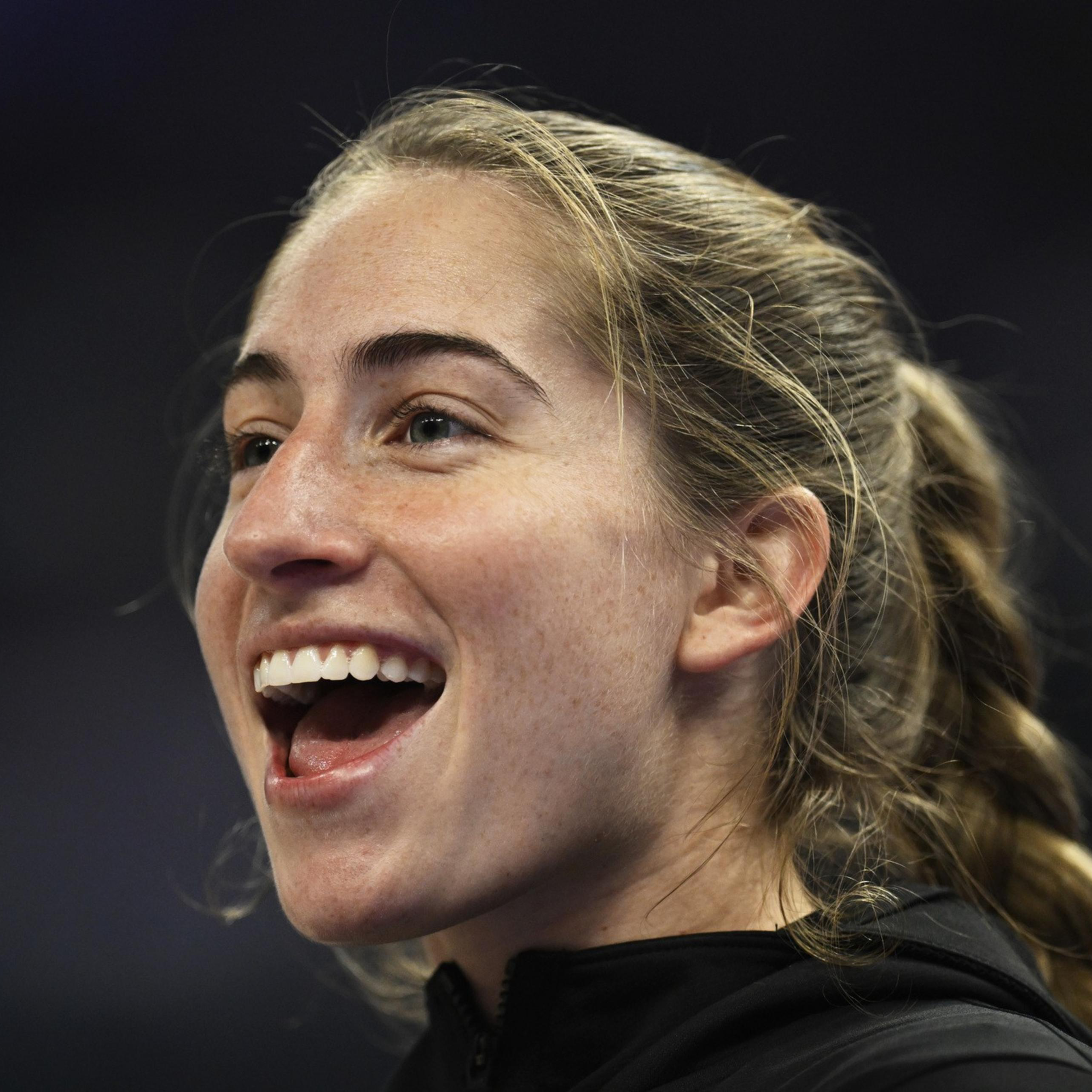 A young woman with blonde hair in a braid is smiling widely, appearing happy and excited, against a dark blurred background.