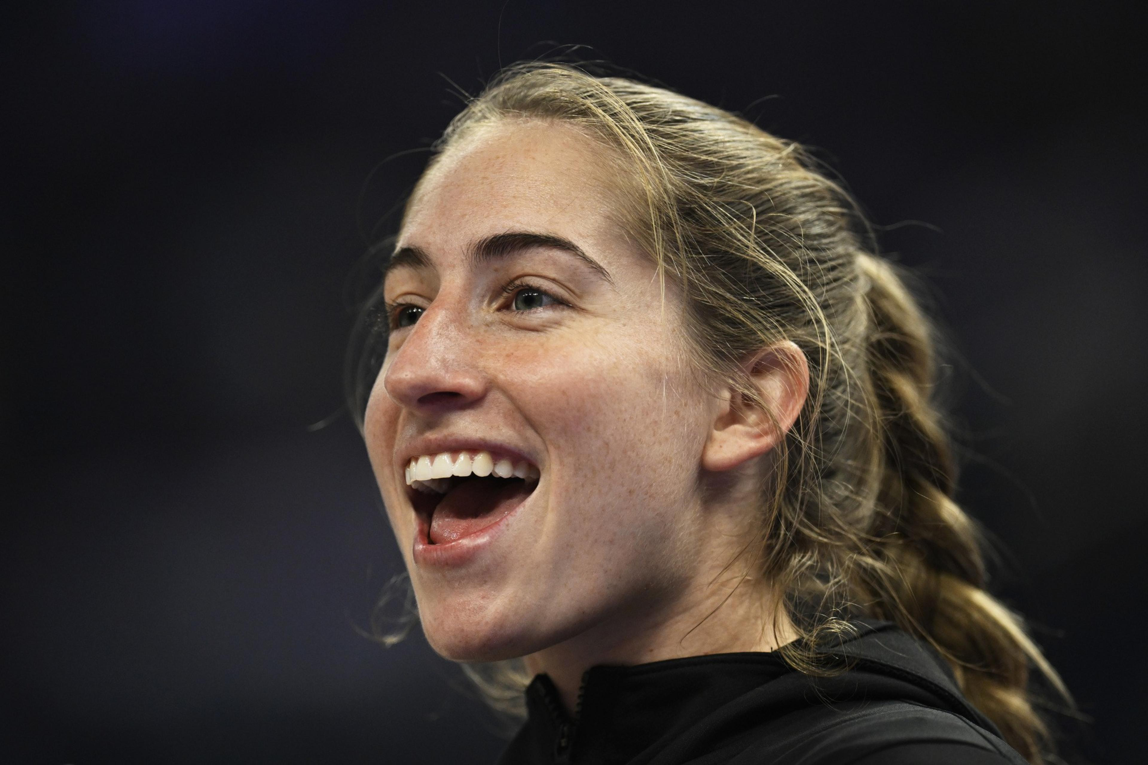 A young woman with blonde hair in a braid is smiling widely, appearing happy and excited, against a dark blurred background.