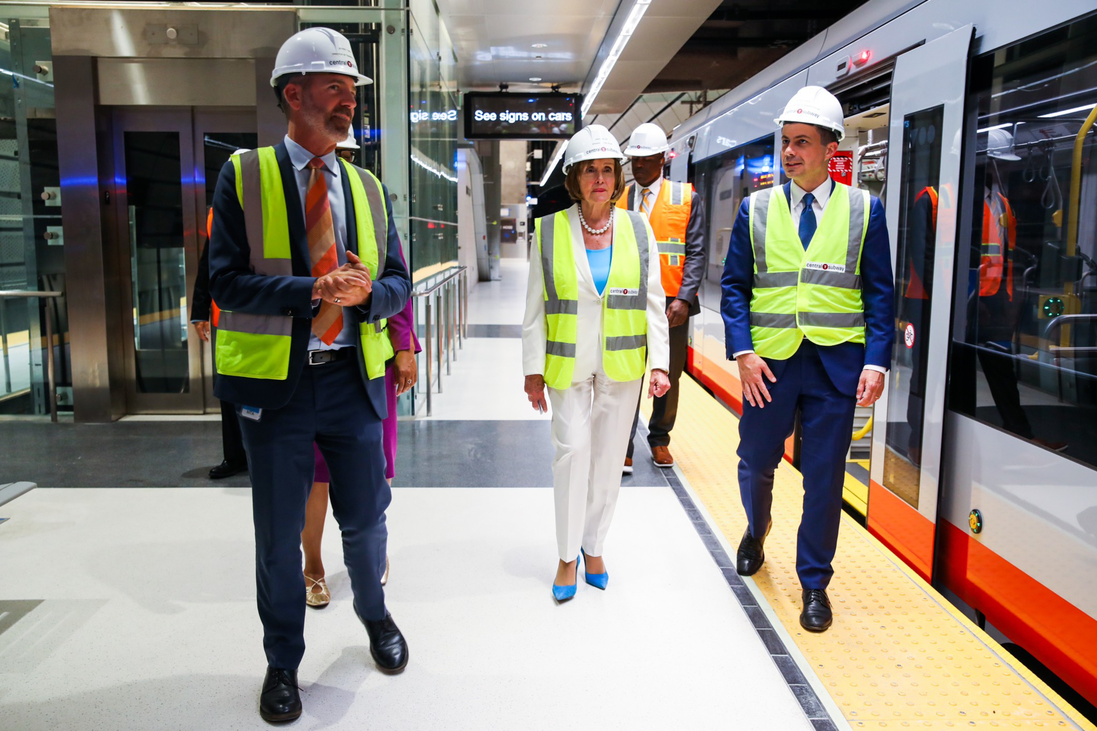 Three people wearing safety vests and hard hats walk along a train platform next to a stationary train inside a modern station.