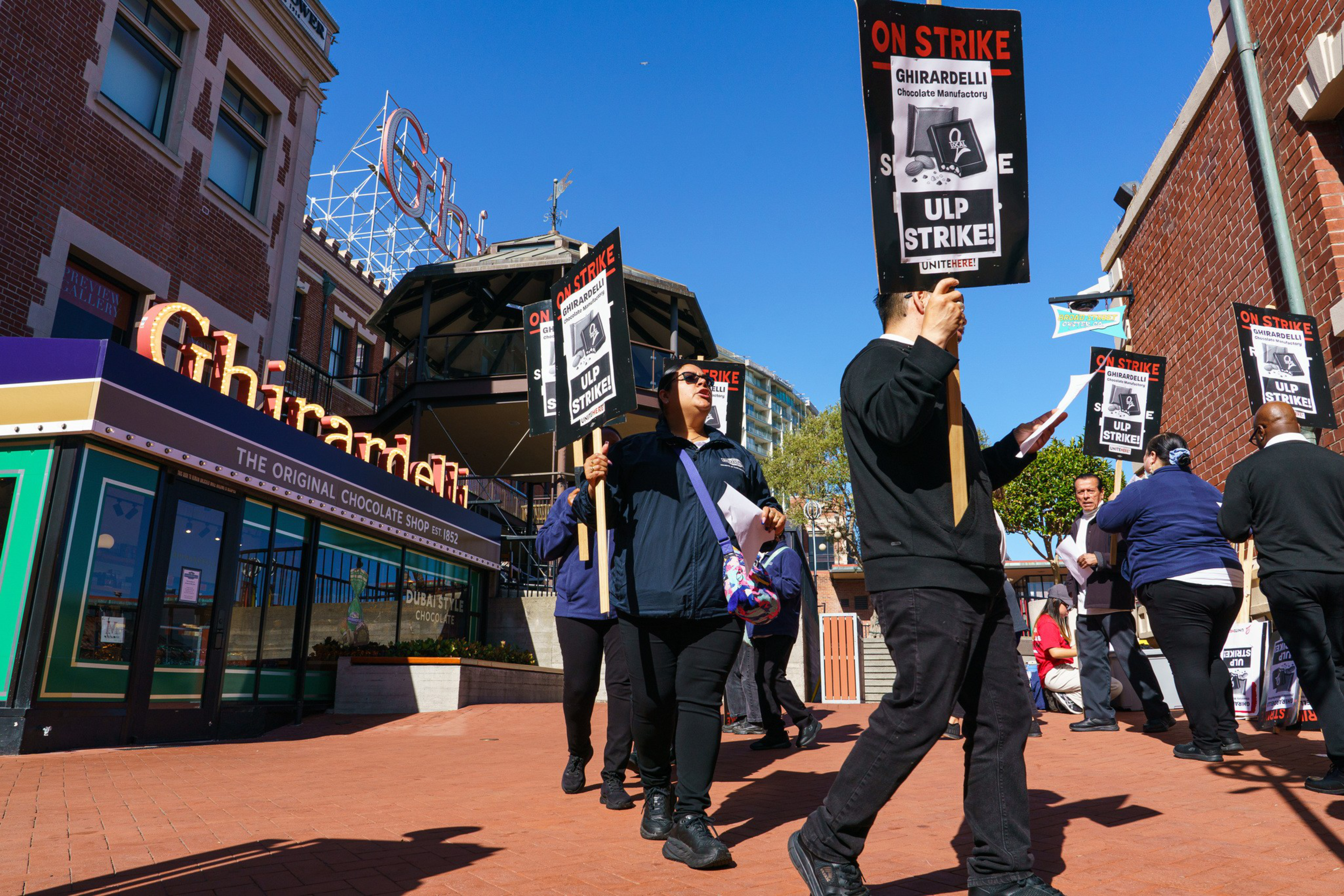 People hold signs reading "ON STRIKE" and "ULP STRIKE!" outside the Ghirardelli chocolate shop during a labor protest on a sunny day.
