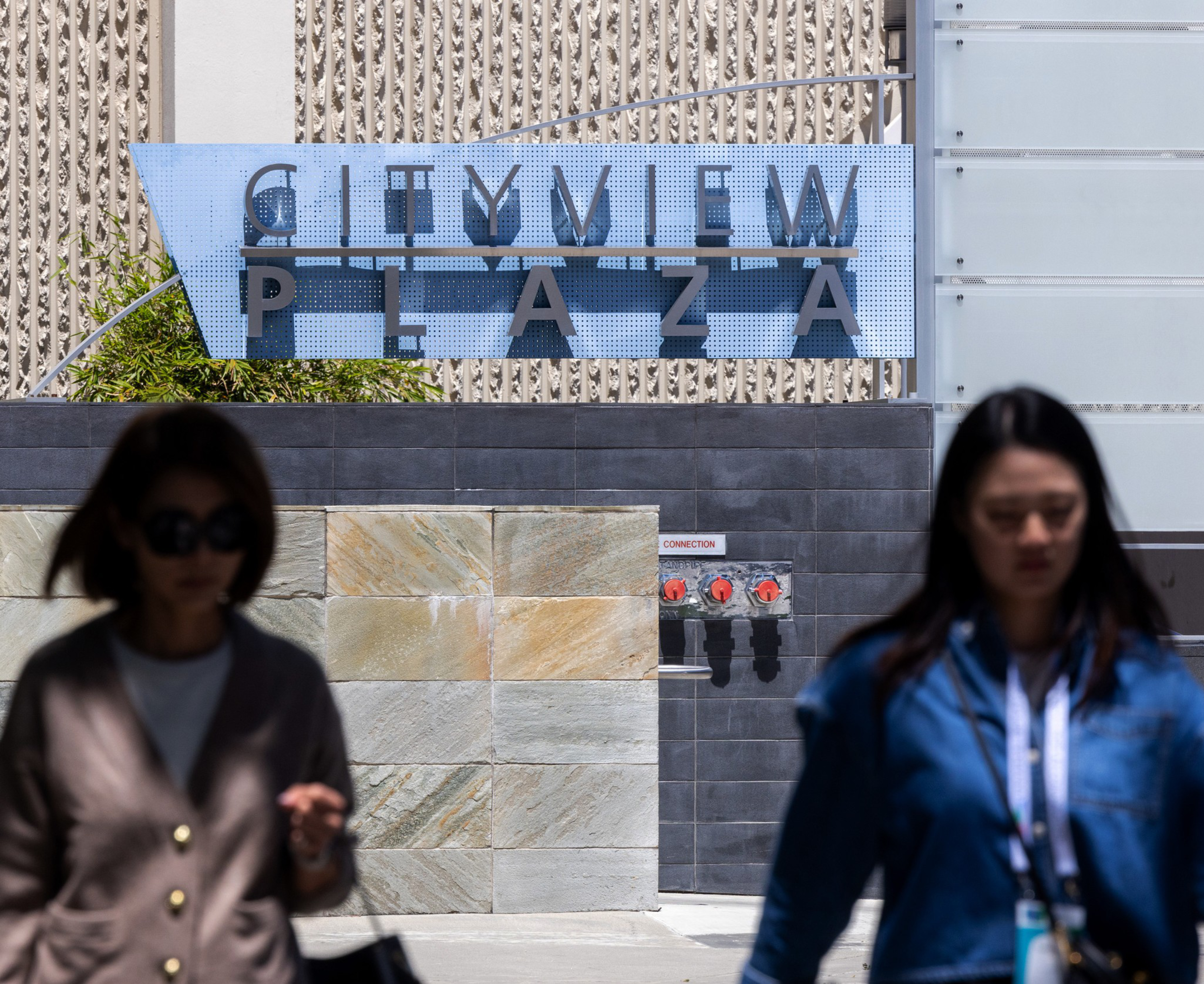 Two people wearing sunglasses walk past a building with a sign that reads &quot;City View Plaza.&quot; The background features textured walls and red pipe connections.