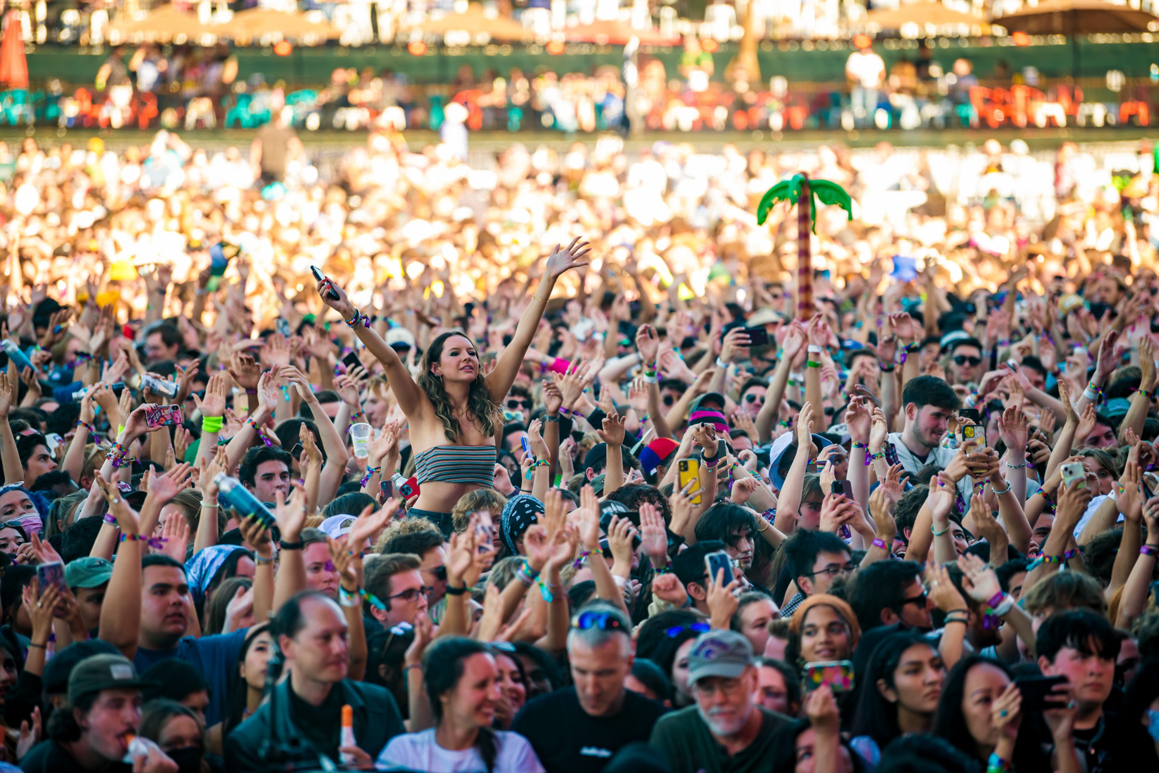 A large crowd at a music festival raises their hands, with a smiling woman on someone's shoulders and colorful accessories like an inflatable palm tree visible.