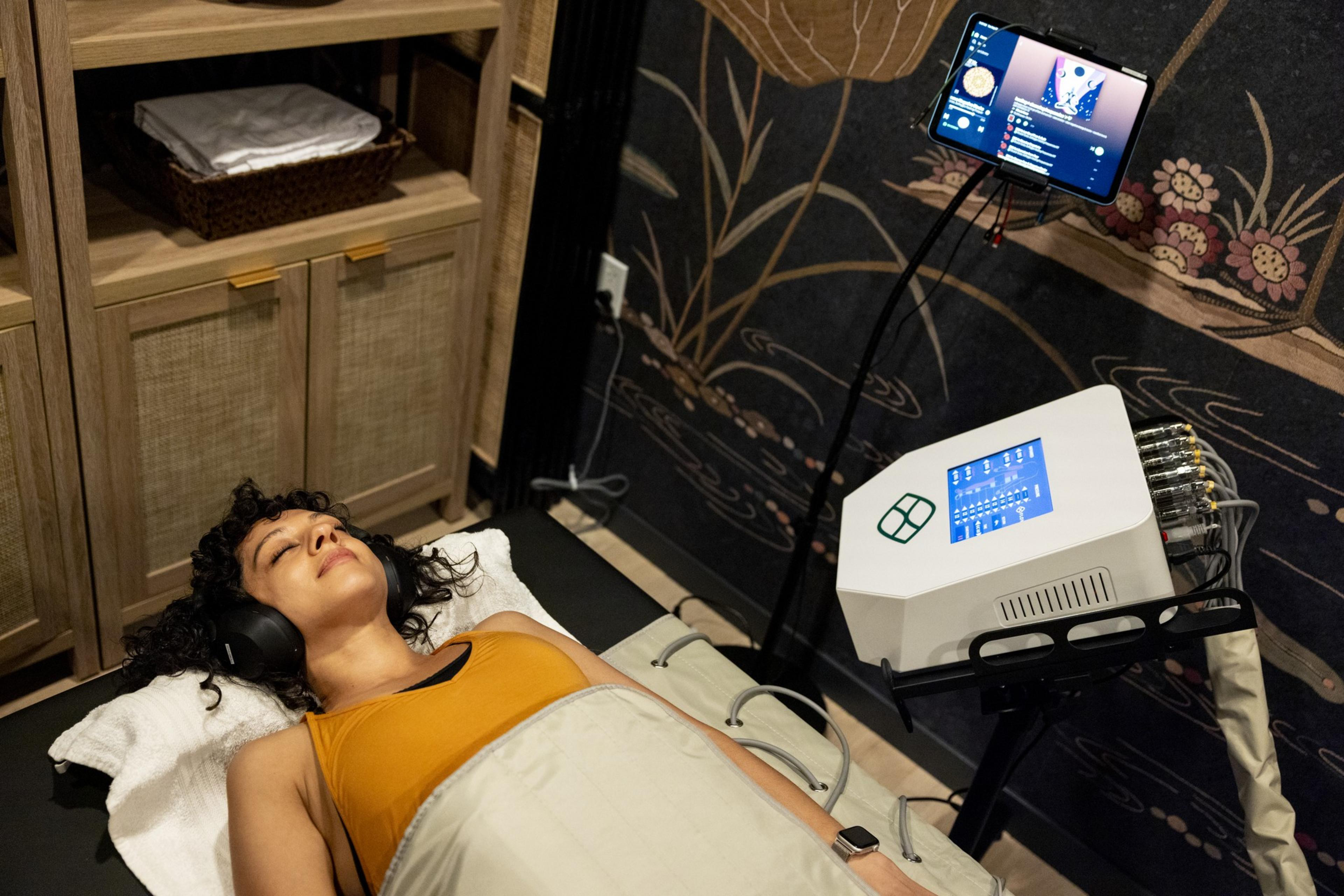 A woman lies on a padded table, wearing headphones and a yellow tank top. She appears relaxed with a machine and screen next to her. The room has wood cabinets and a decorative wall.