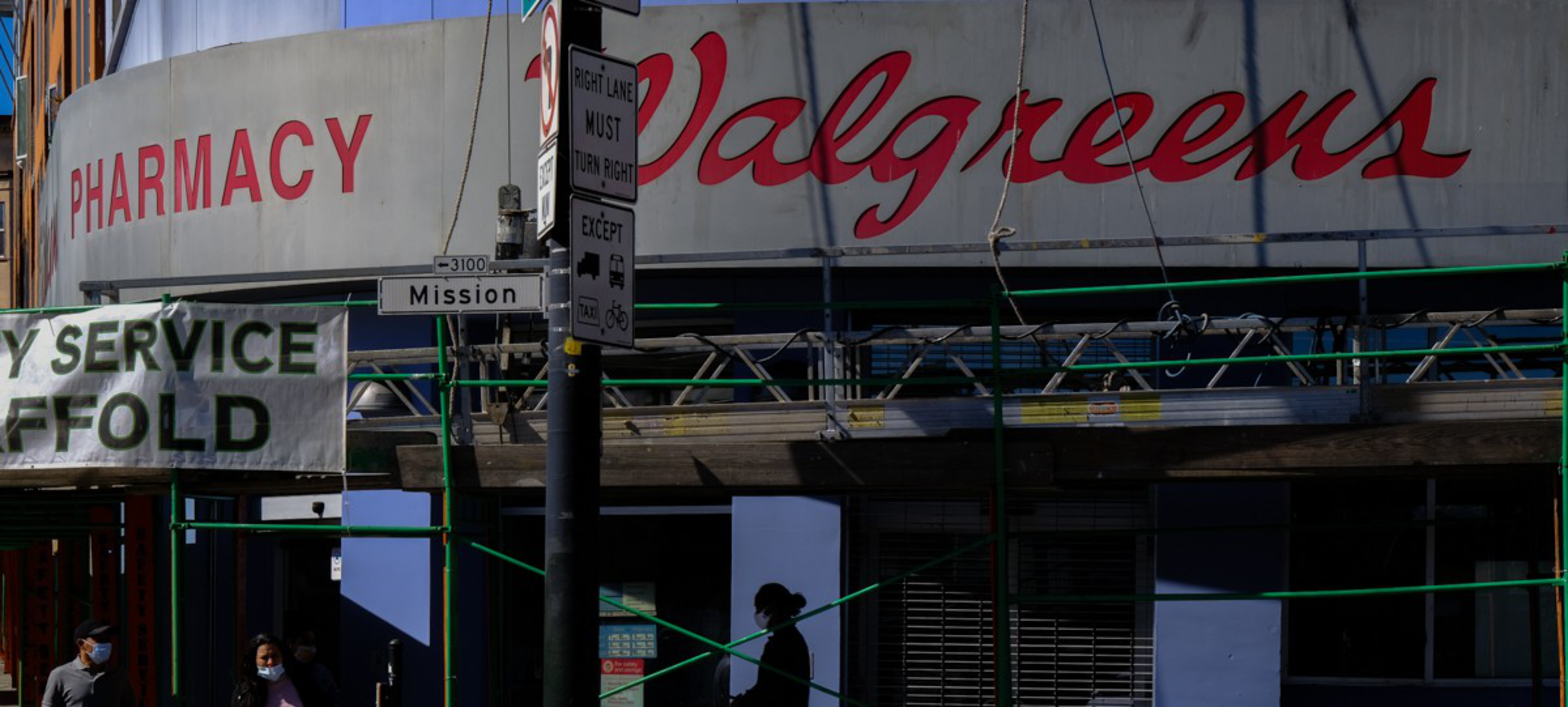 A Walgreens pharmacy storefront with scaffolding and a street sign reading Mission stands under a clear sky, with a few people walking by wearing masks.