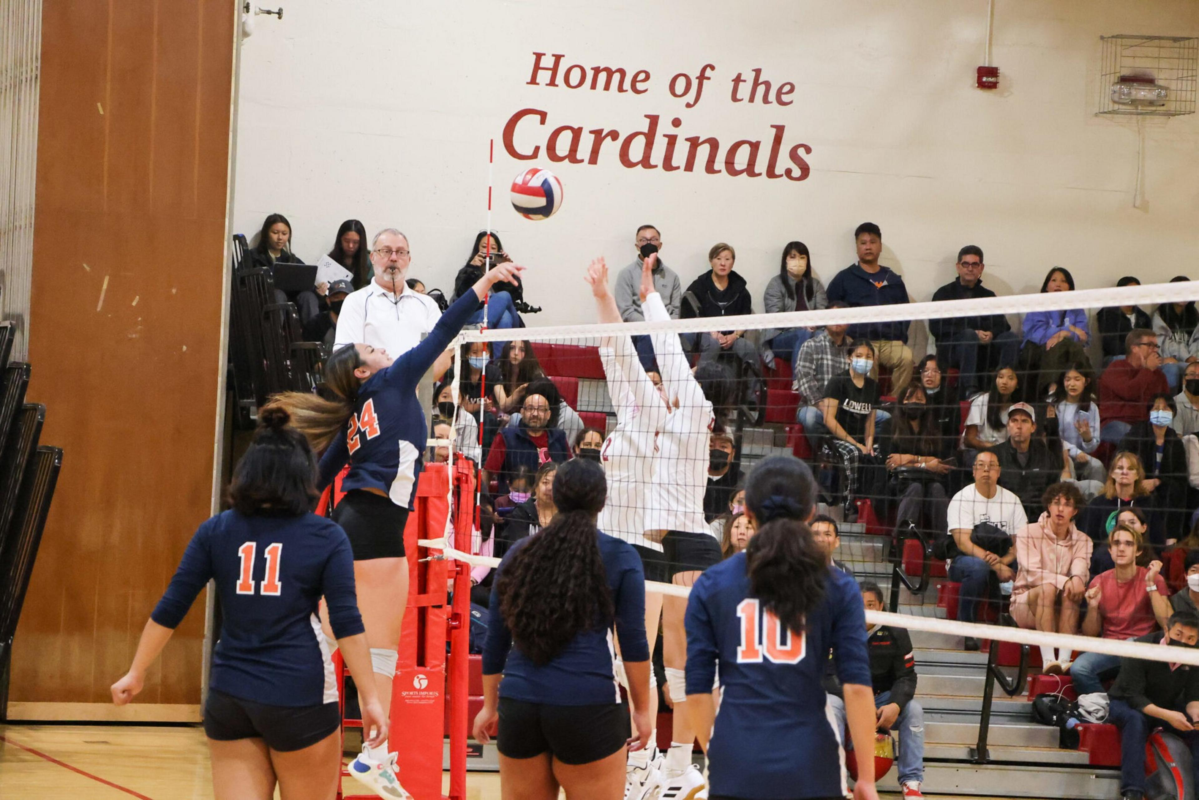 A volleyball player in a navy jersey jumps to spike the ball as an opponent in white jumps to block, with teammates and spectators watching.