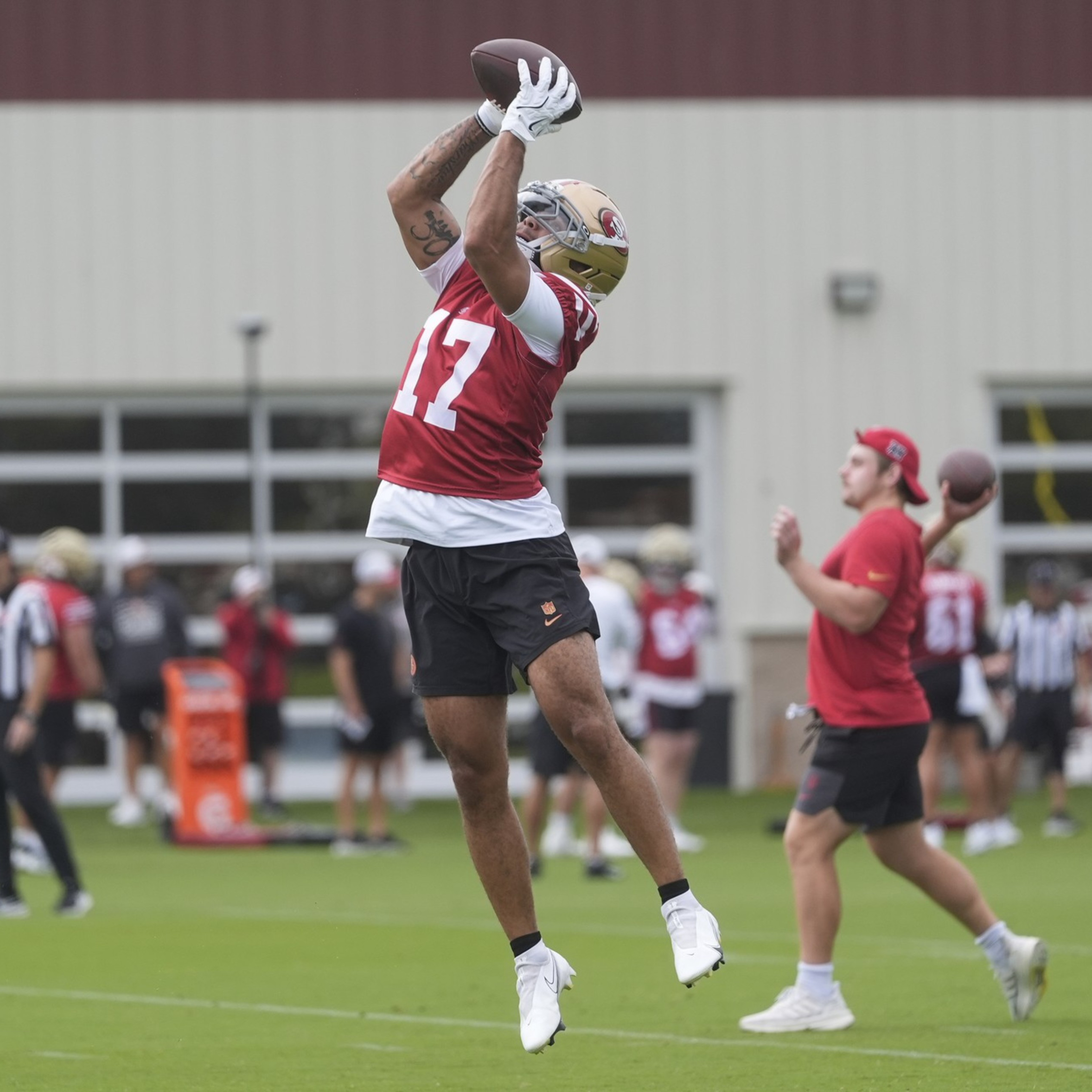 A football player in a red jersey leaps to catch a pass, while others practice in the background on a field.