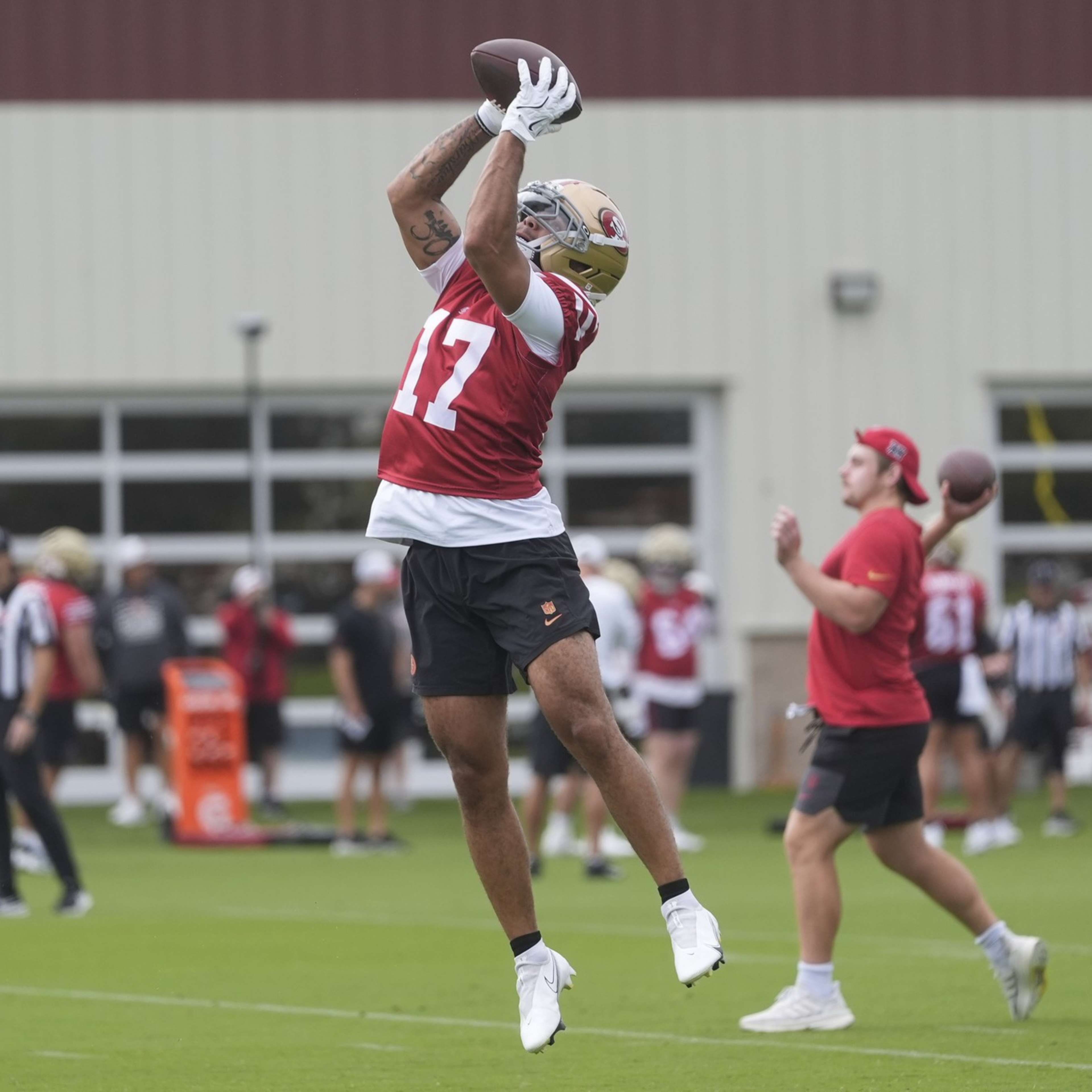 A football player in a red jersey leaps to catch a pass, while others practice in the background on a field.