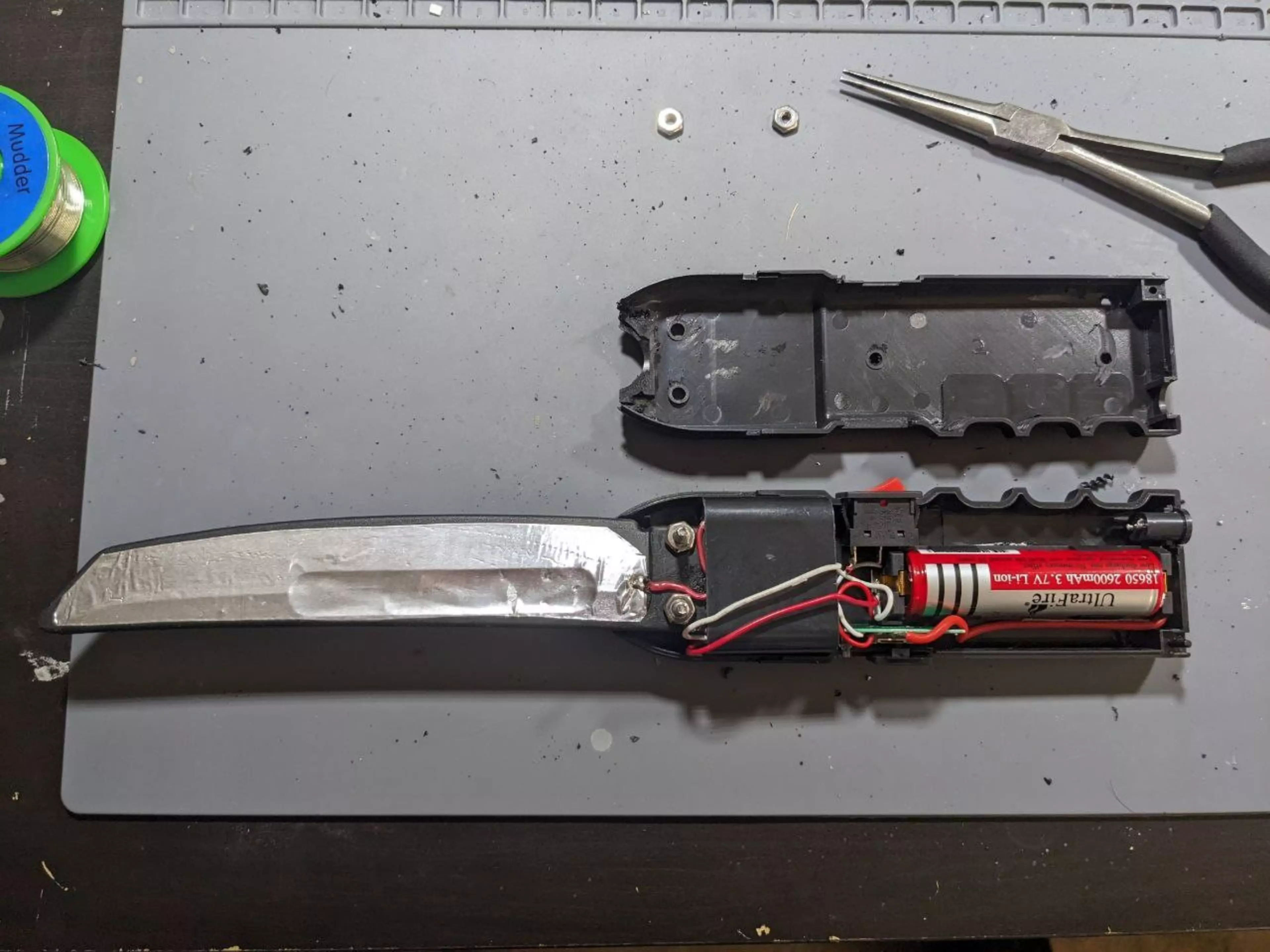 A disassembled electronic knife with battery, wiring, and internal components laid out on a gray mat, accompanied by small hardware and pliers.