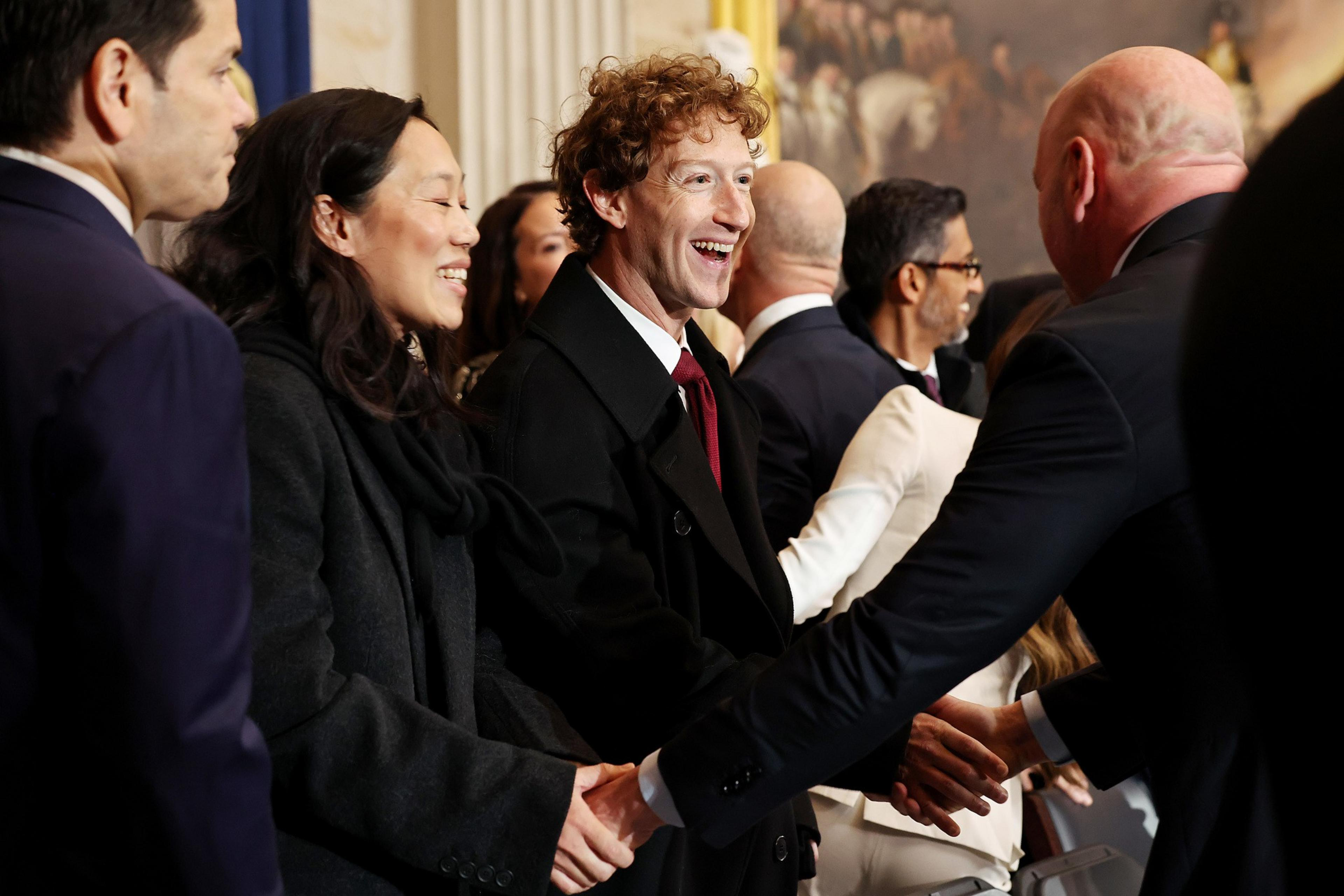 A group of people in formal attire are gathered, smiling and shaking hands. The setting appears to be a formal event or gathering.