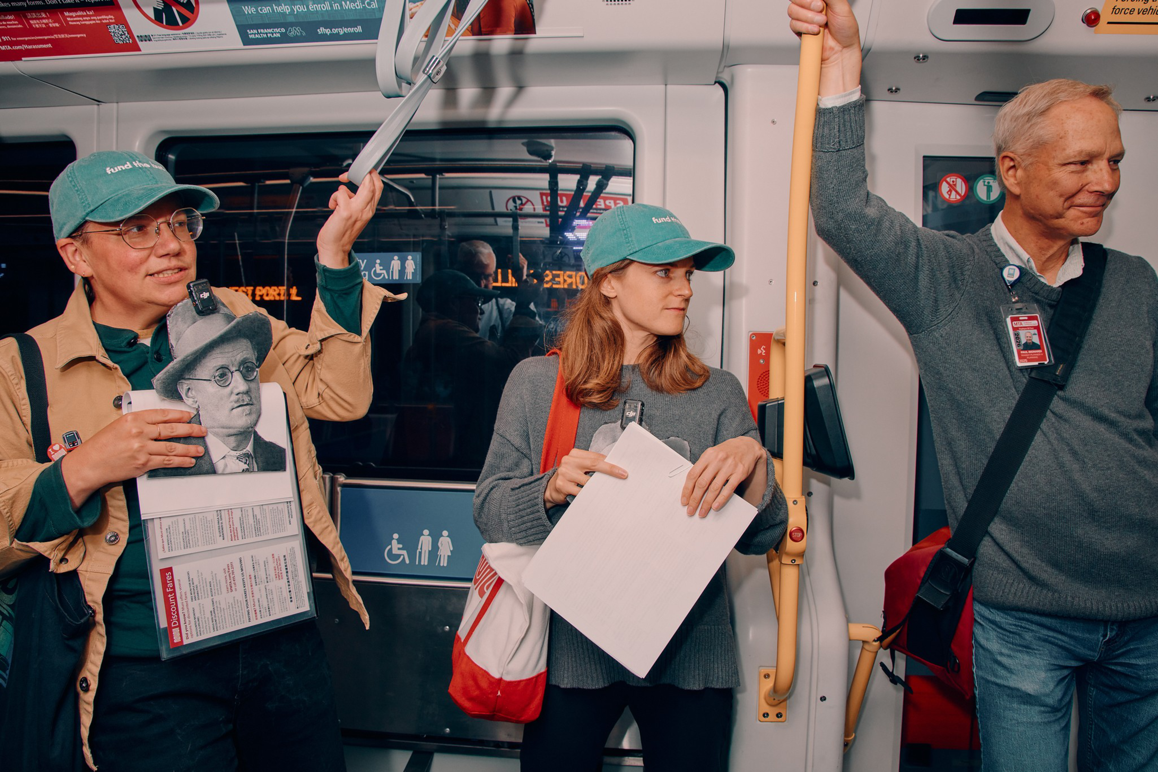 Three people stand inside a public transit vehicle, two holding papers and one holding a stick for support, all wearing casual clothing and bags.