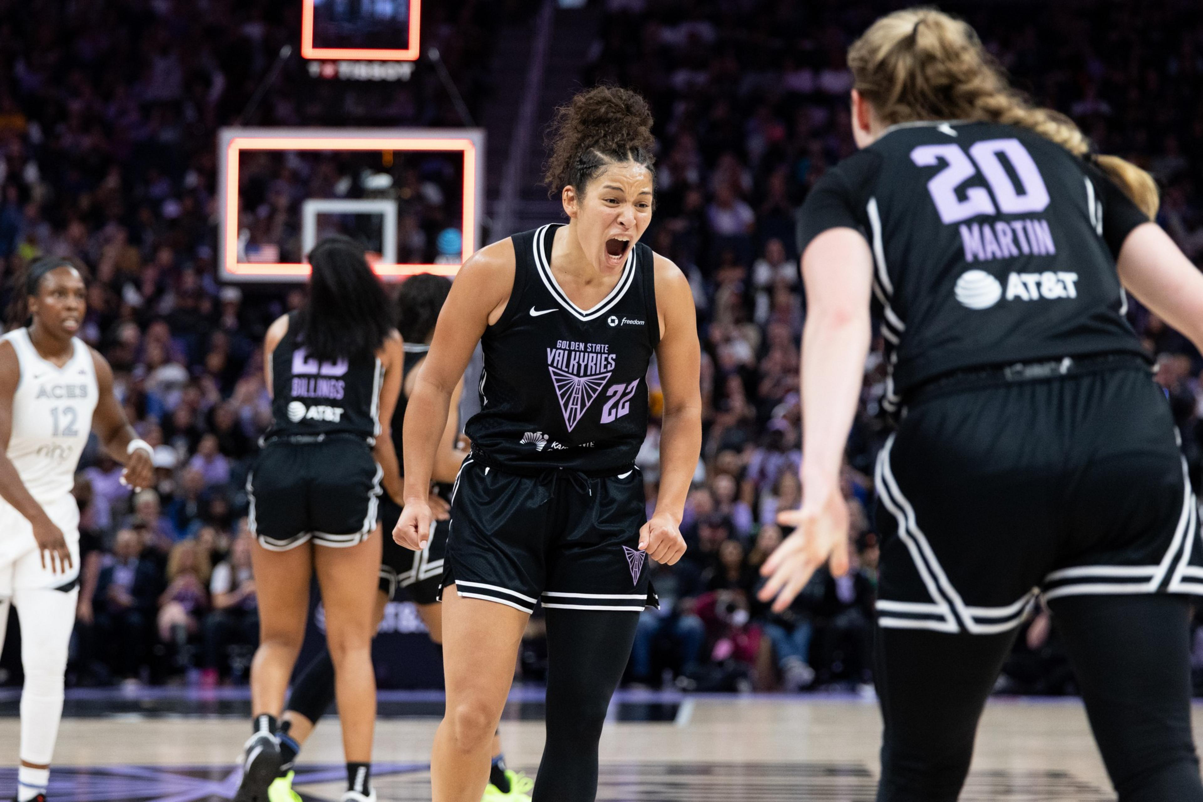 Two basketball players in black jerseys are on the court, one appears to be cheering or shouting. Another player in a white jersey is in the background.