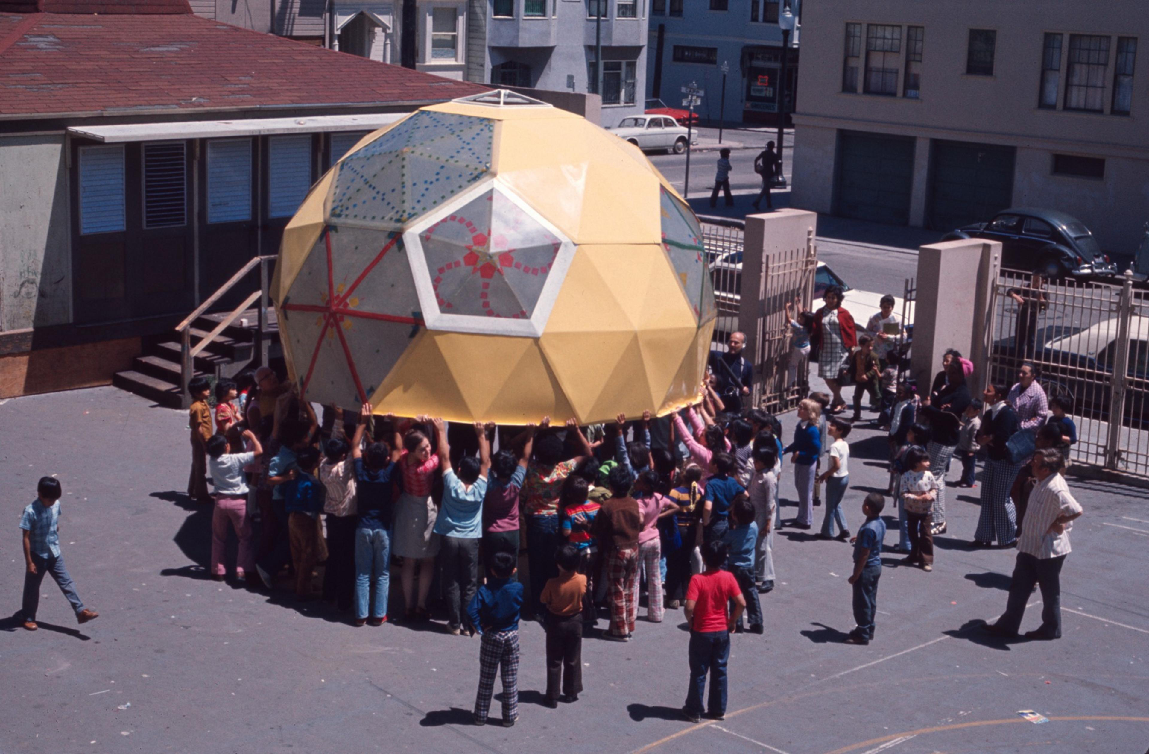 A group of children are lifting a large, colorful geodesic dome in a schoolyard. Nearby, adults and more children are watching and standing around.