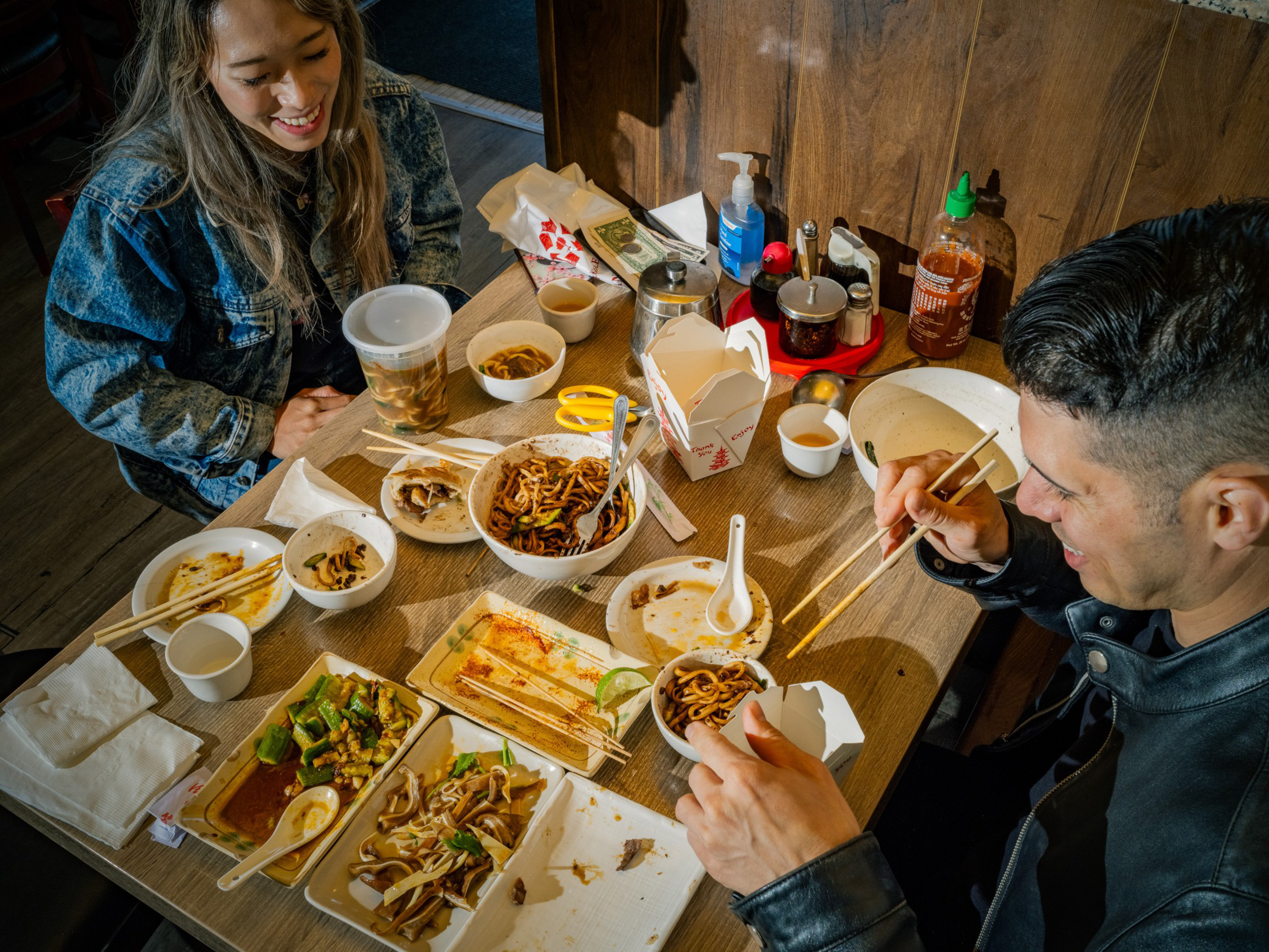 Two people enjoy a meal with various Asian dishes, chopsticks, sauce bottles, and takeout containers on a wooden table.