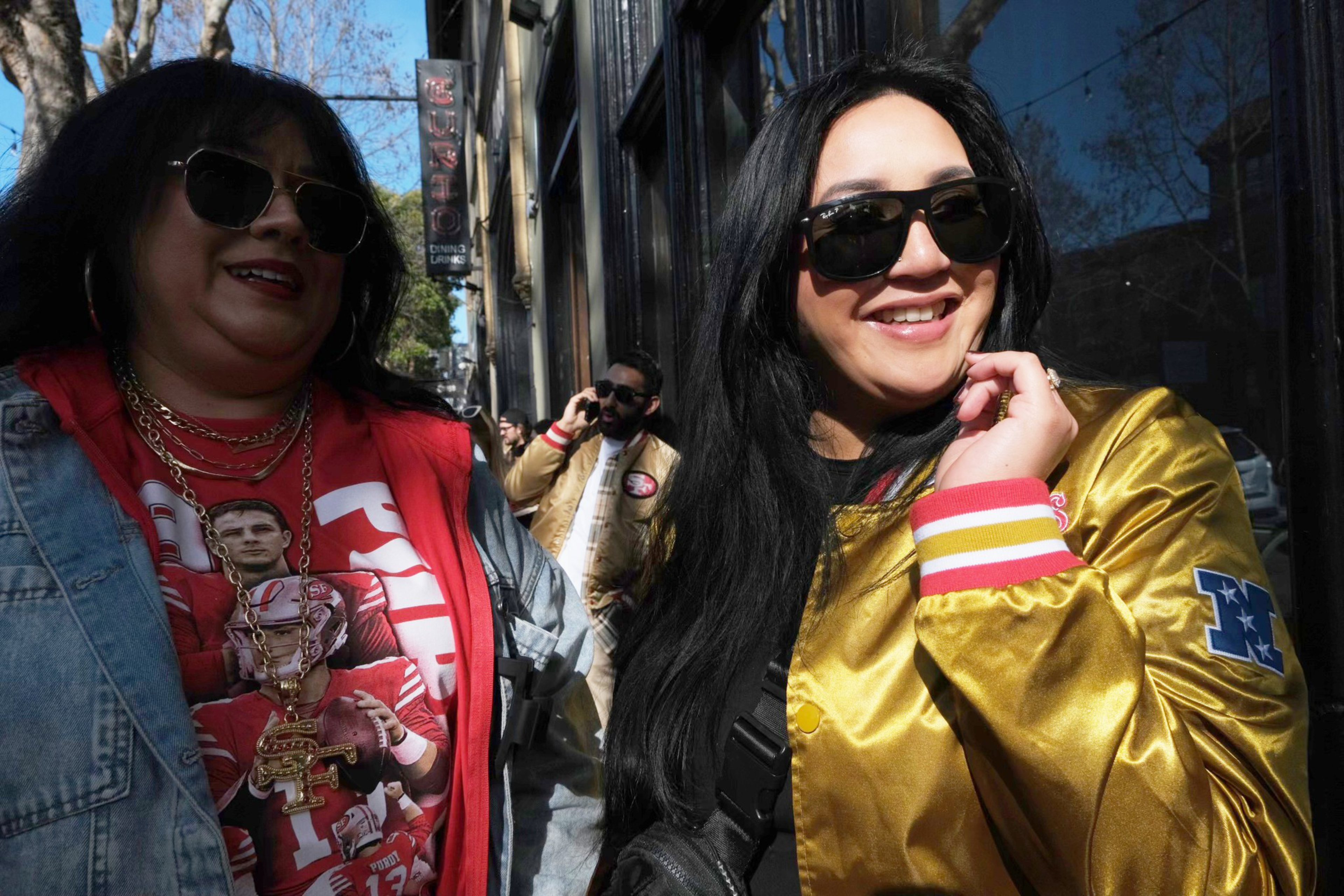 Two women smiling in the sun, wearing sunglasses and sports-themed attire, with a man in background on phone.