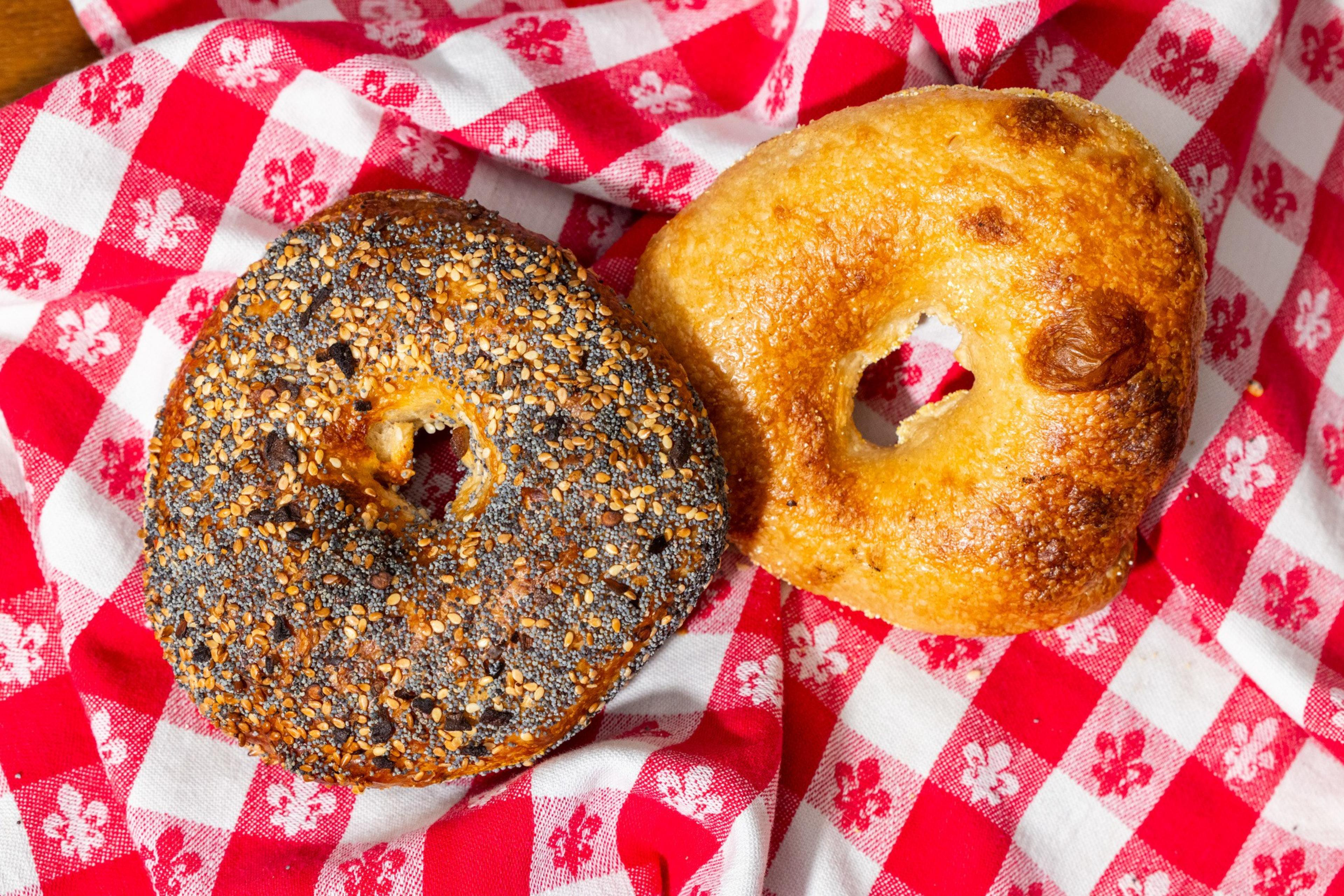 Two bagels rest on a red and white checkered cloth. One is seeded, featuring poppy and sesame seeds, while the other is plain and golden-brown.