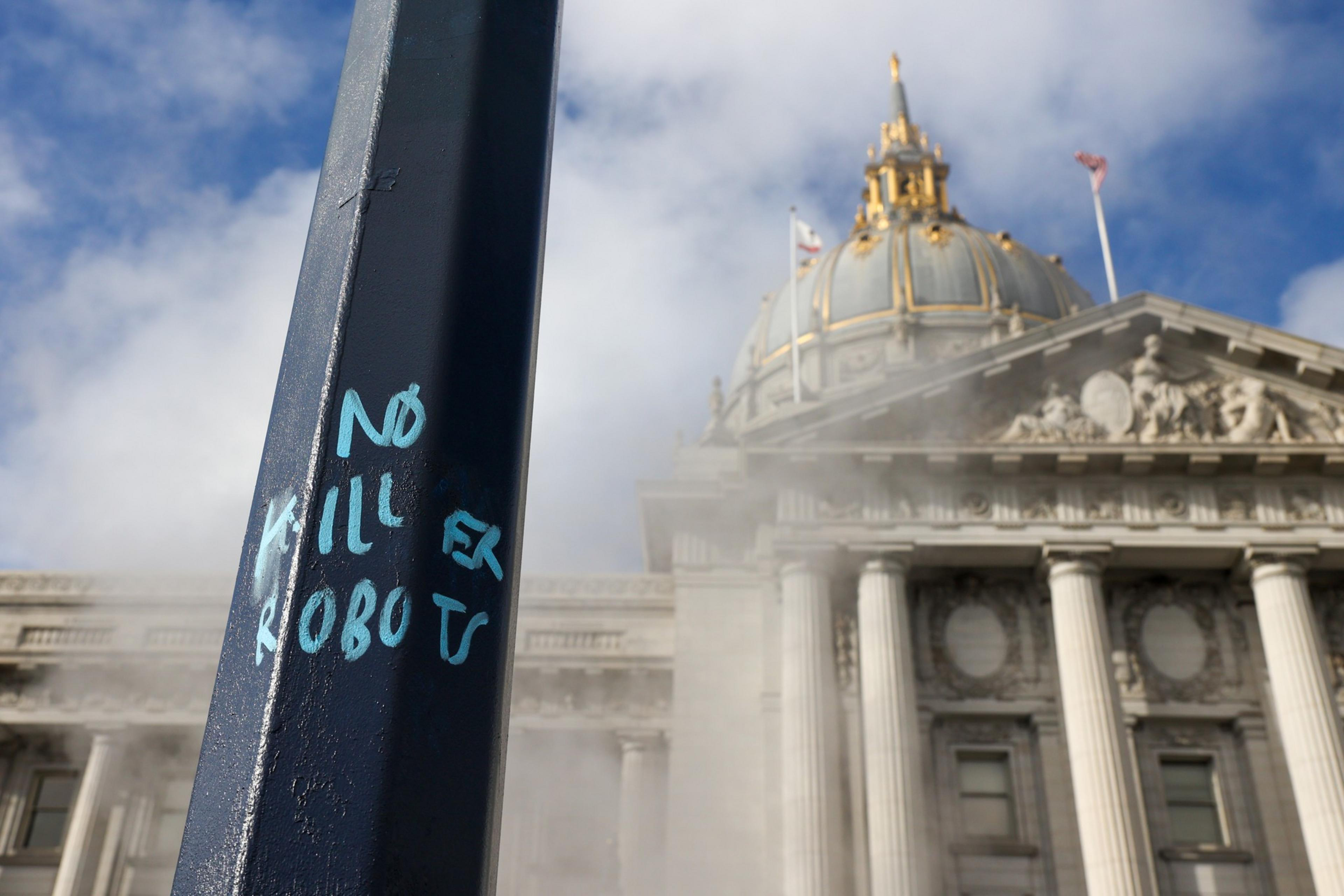 A black pole with blue graffiti reading "No Killer Robots" stands in front of a classical building with columns and a gold-domed roof under a partly cloudy sky.