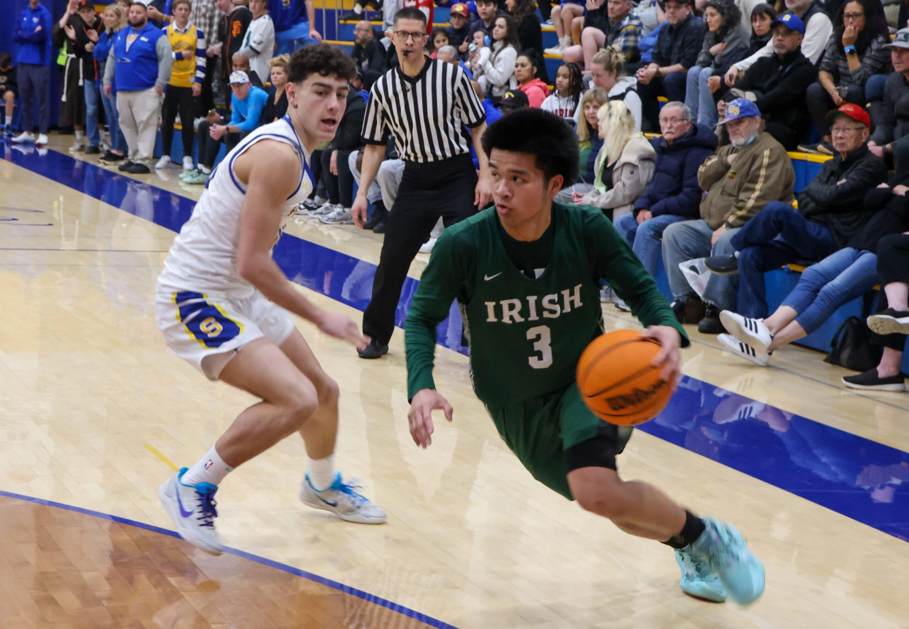 A basketball player in a green "Irish" jersey dribbles past a defender in white while a referee and crowd watch from the sidelines.