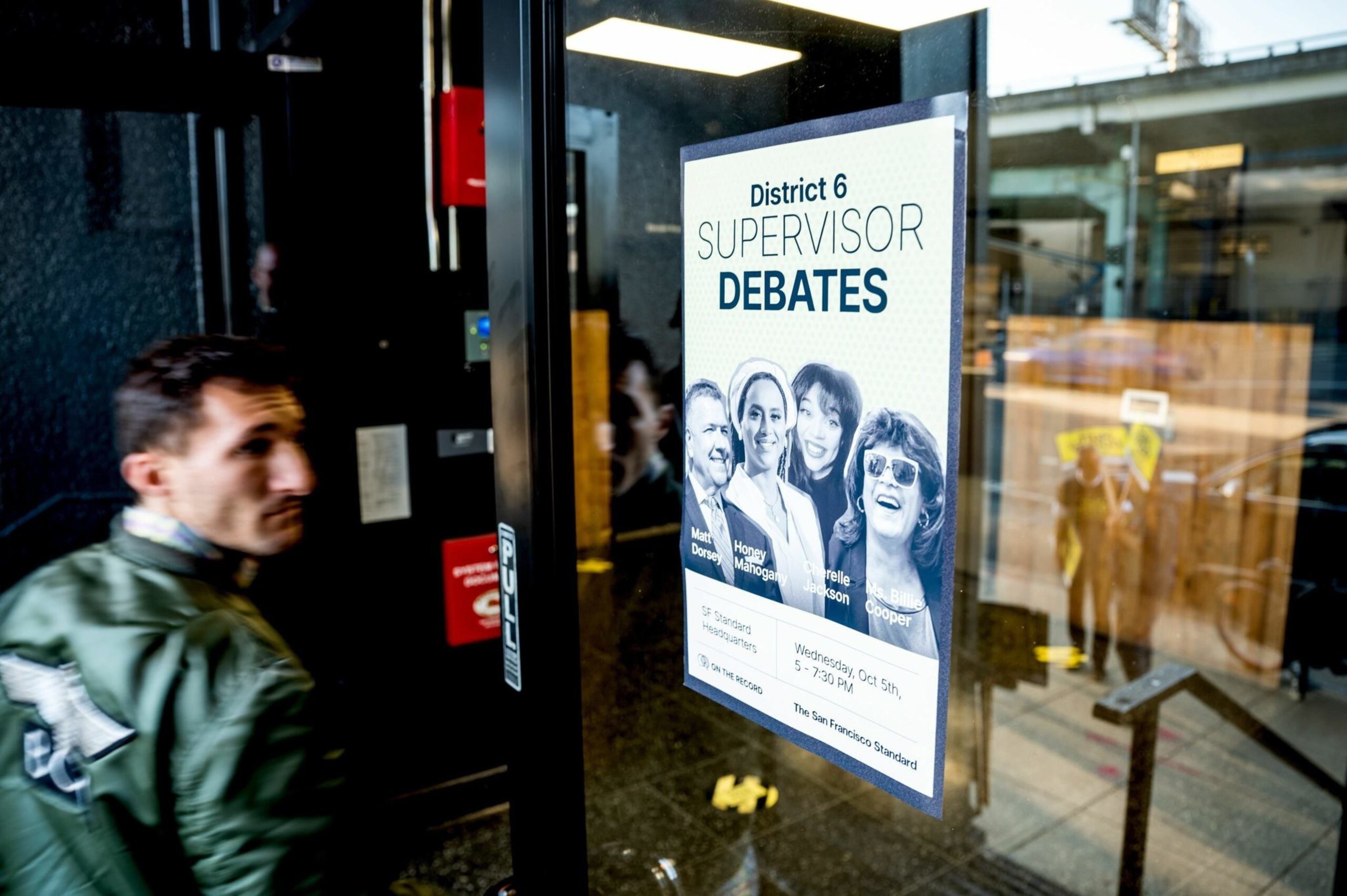 A poster on a glass door advertises the District 6 Supervisor Debates featuring four candidates, with a man walking past it inside a building.