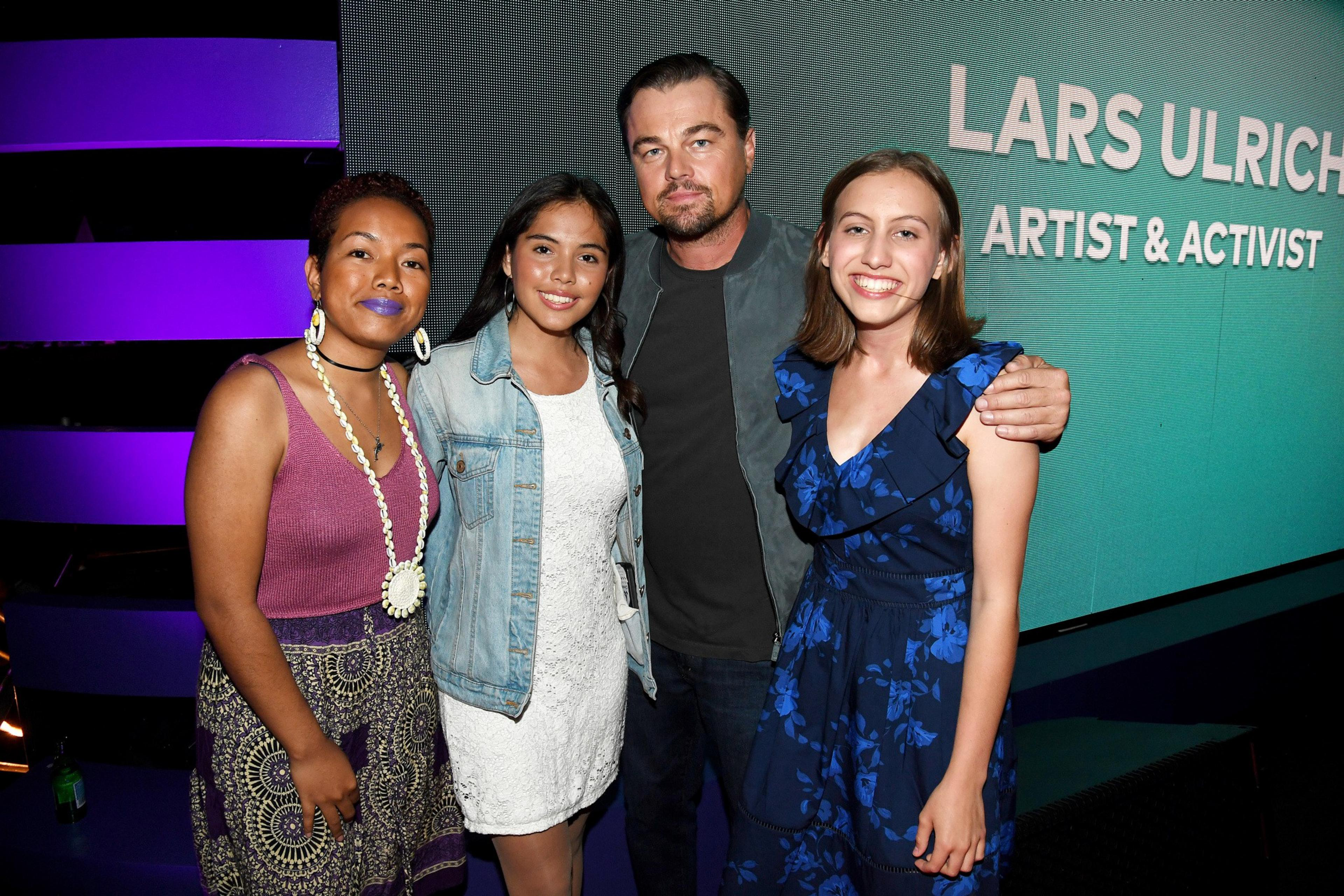 A man and three women smile for a photo; the man wears a black jacket and the women wear colorful dresses. The background features the text &quot;Lars Ulrich, Artist &amp; Activist.&quot;