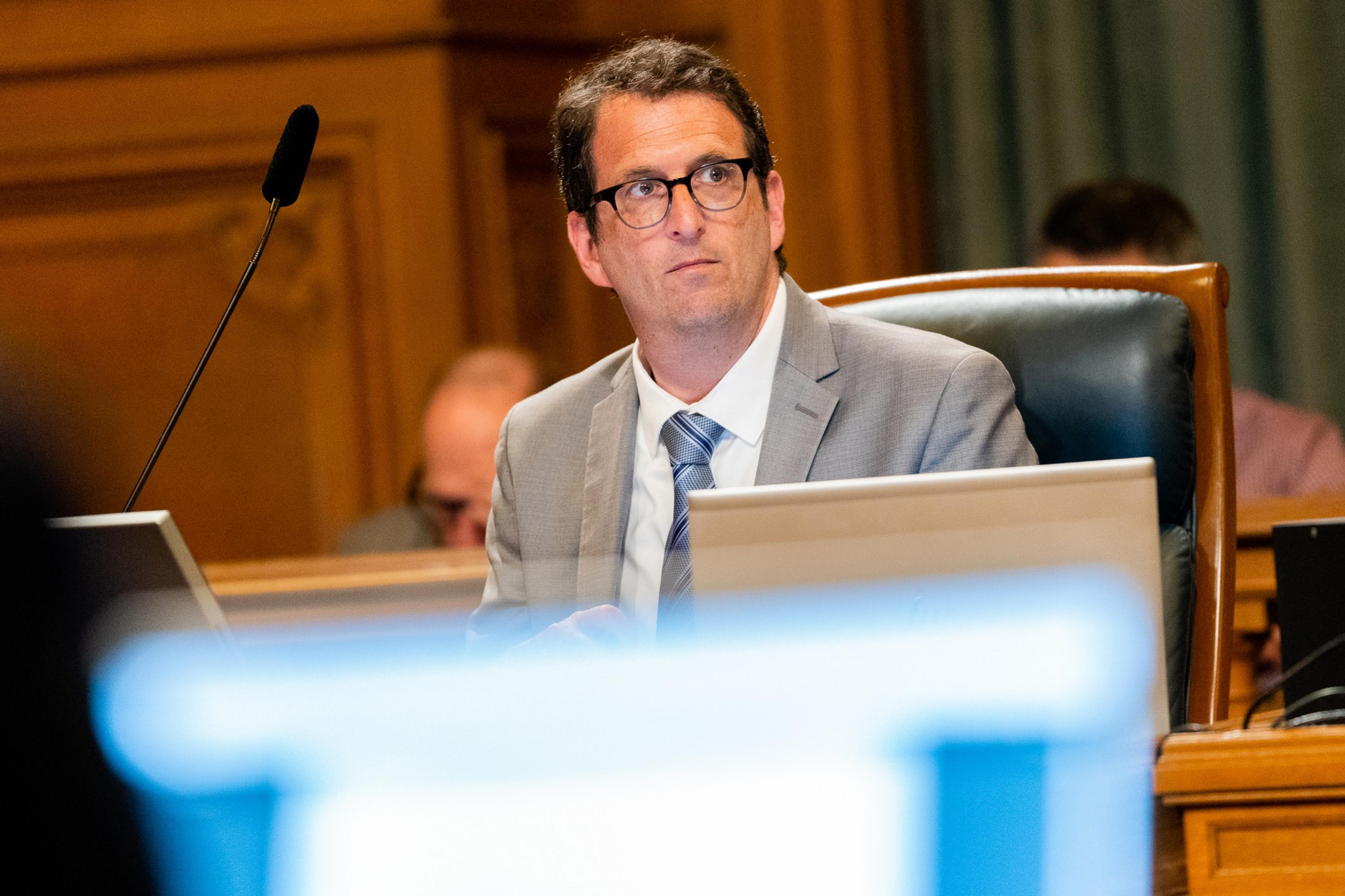 A man in a light gray suit, blue tie and glasses glances to his left.