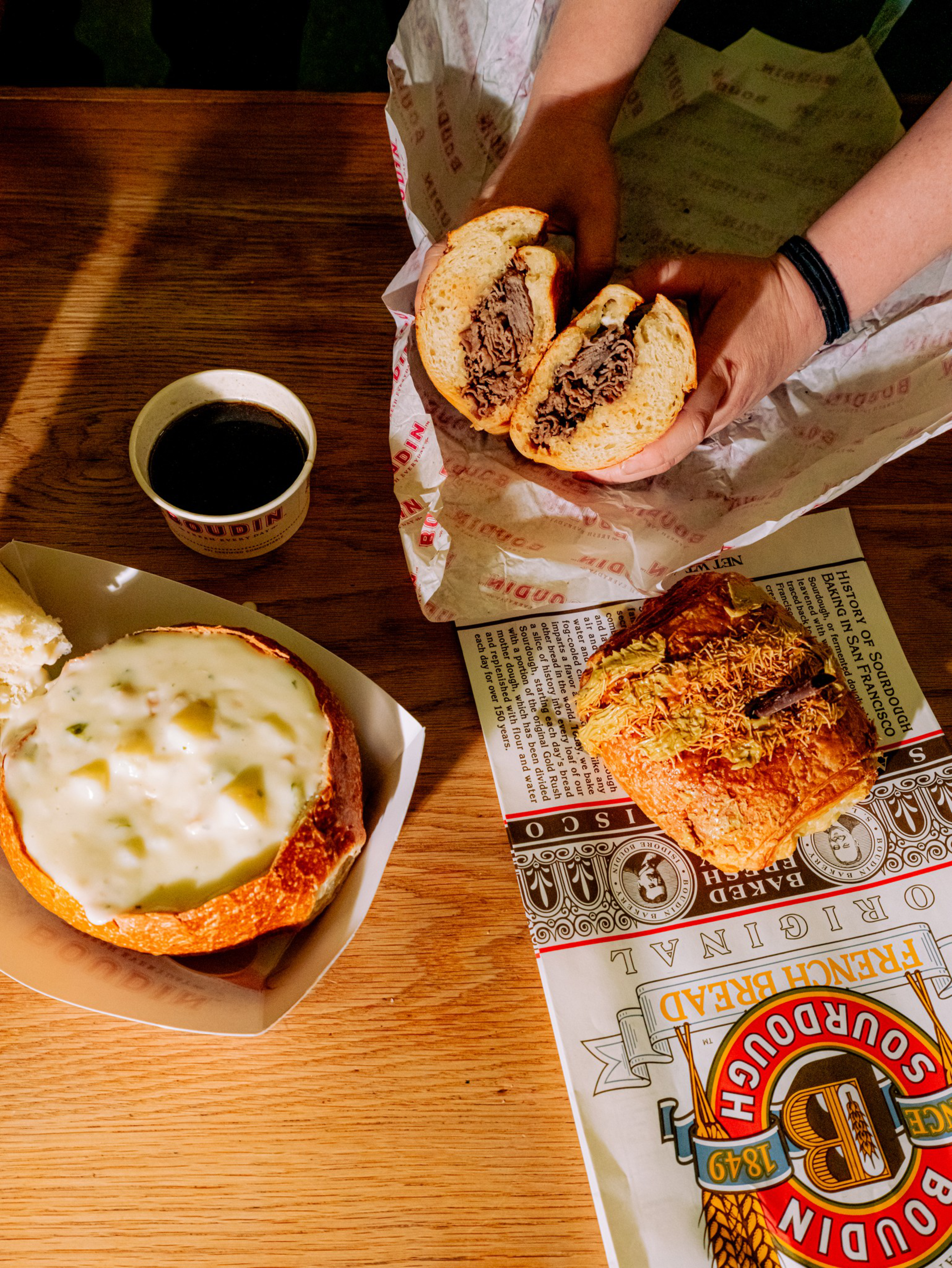 A person holds a sliced roast beef sandwich on parchment paper, next to a bread bowl filled with creamy soup and a cup of black coffee on a wooden table.