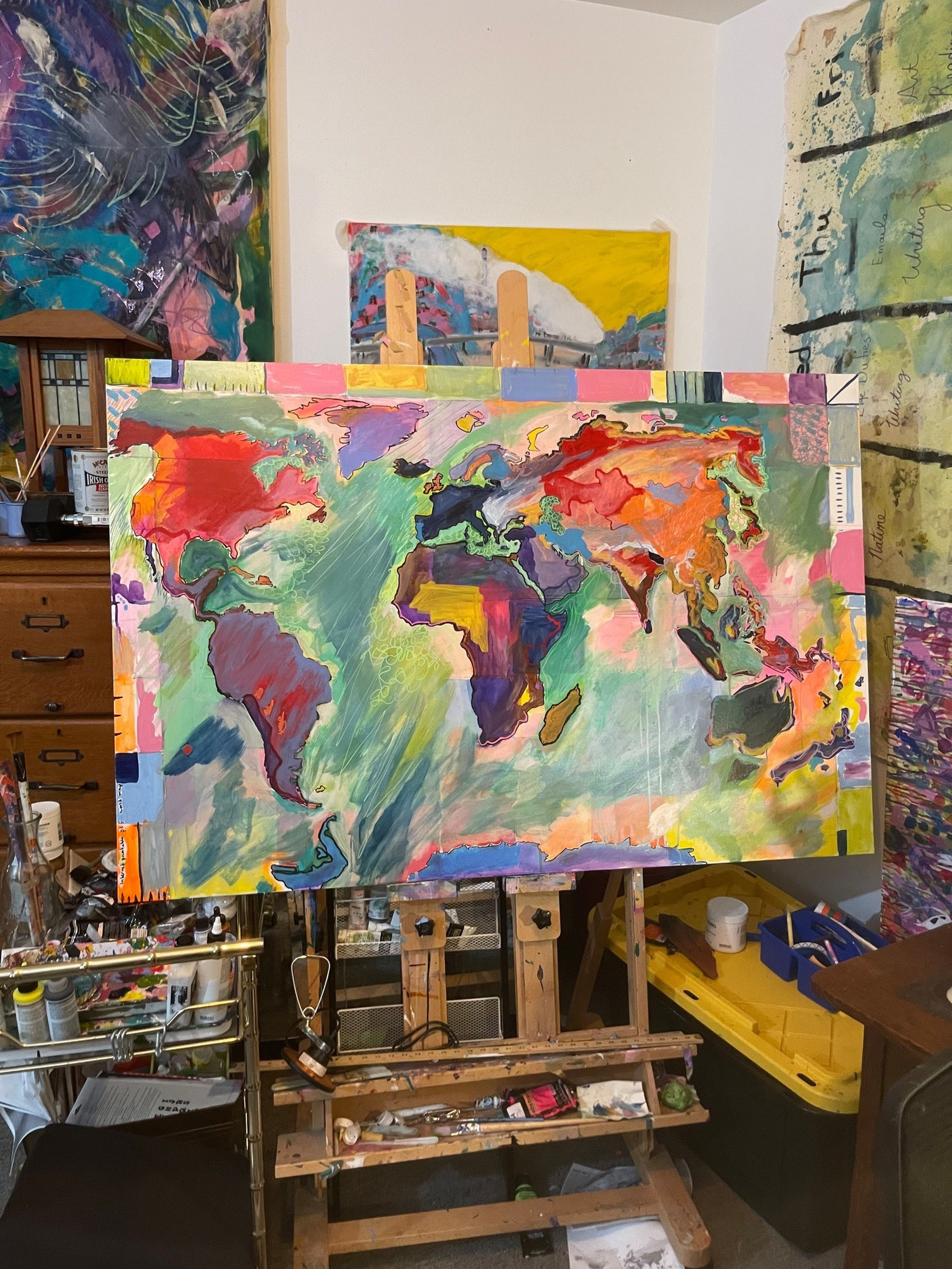 A colorful, abstract world map painting rests on an easel in a cluttered artist’s studio filled with various canvases, paints, and art supplies.
