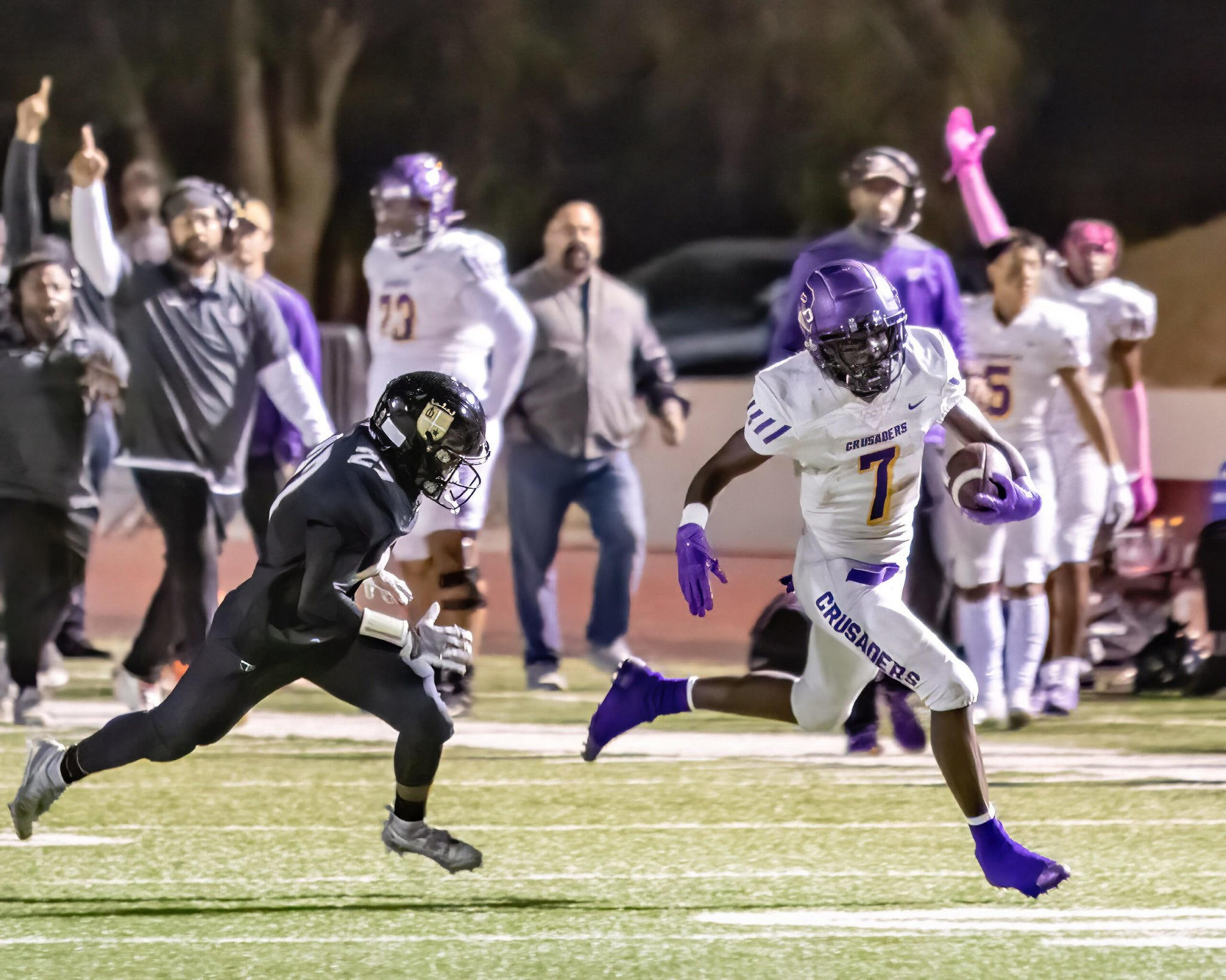 A football player in a white uniform runs with the ball as a player in black chases him, while coaches and teammates watch and react from the sidelines.