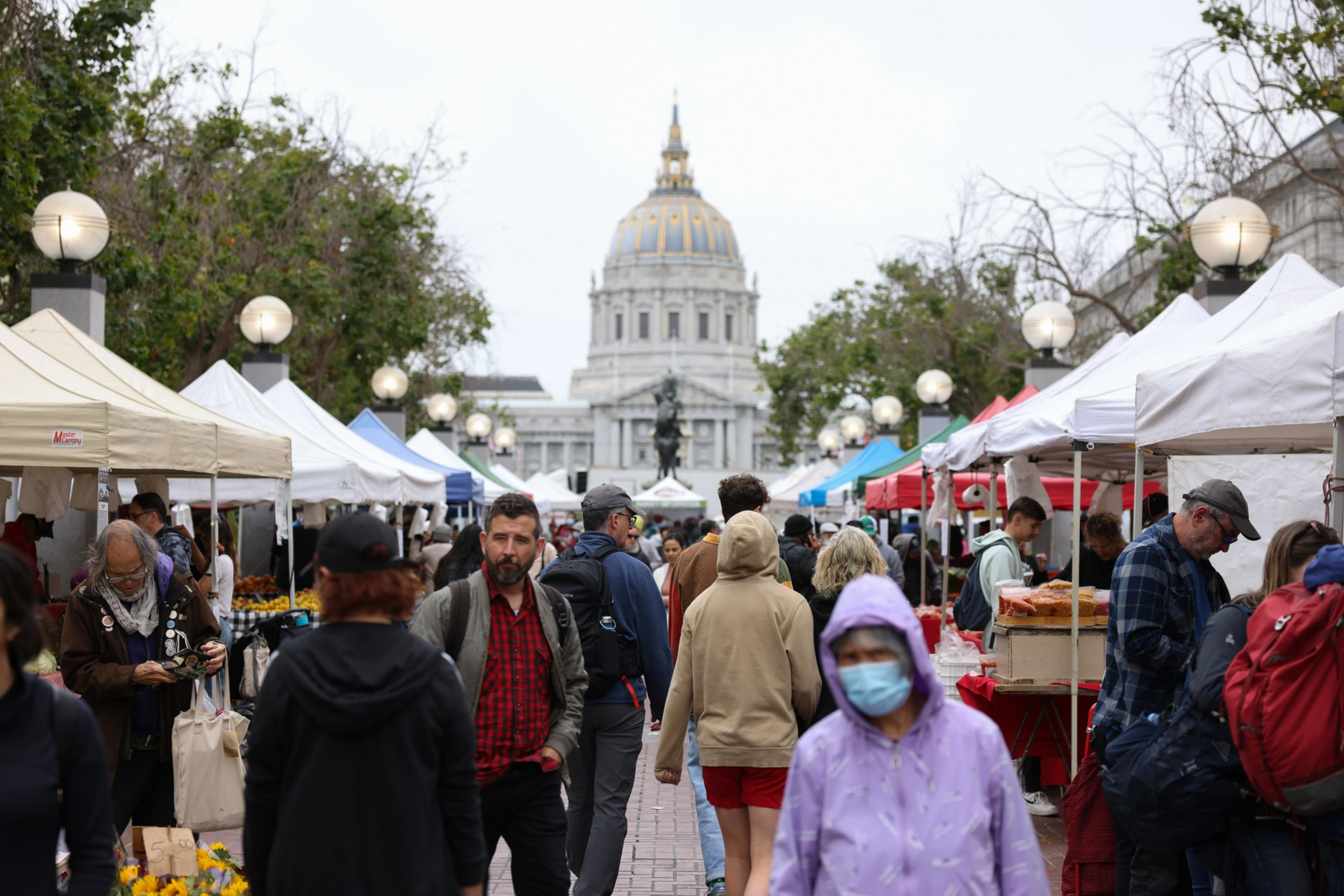 A bustling outdoor market with white tents, people shopping, and a domed building in the background.