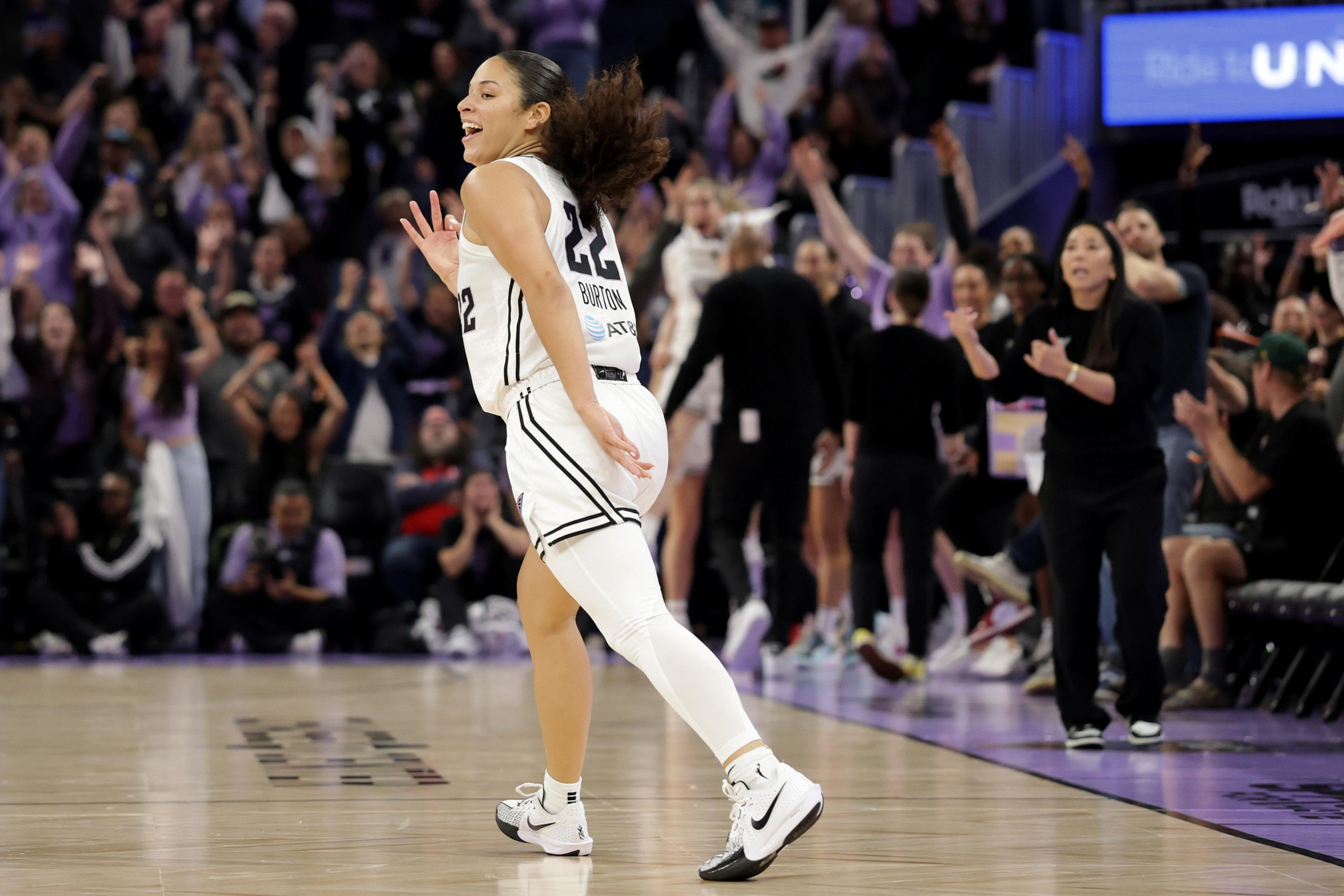 A basketball player in a white uniform celebrates on the court, smiling. The audience and team staff in the background are cheering and applauding.