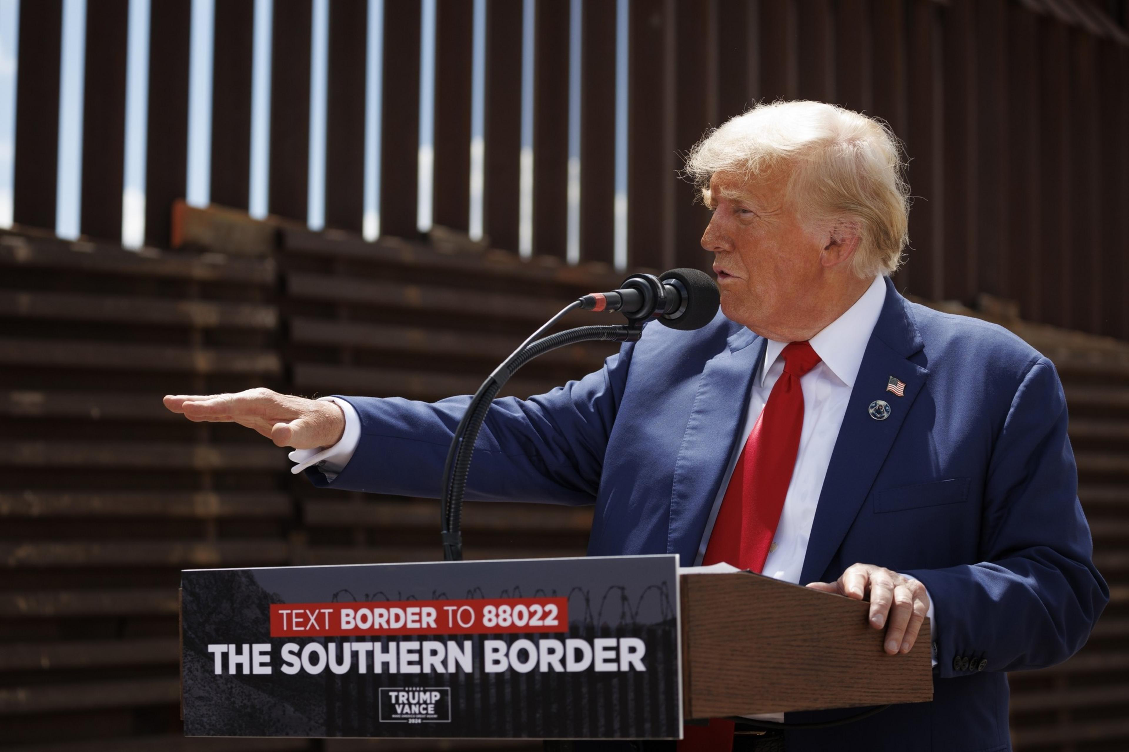 A man in a blue suit and red tie speaks at a podium with &quot;The Southern Border&quot; written on it, in front of a tall metal border fence.